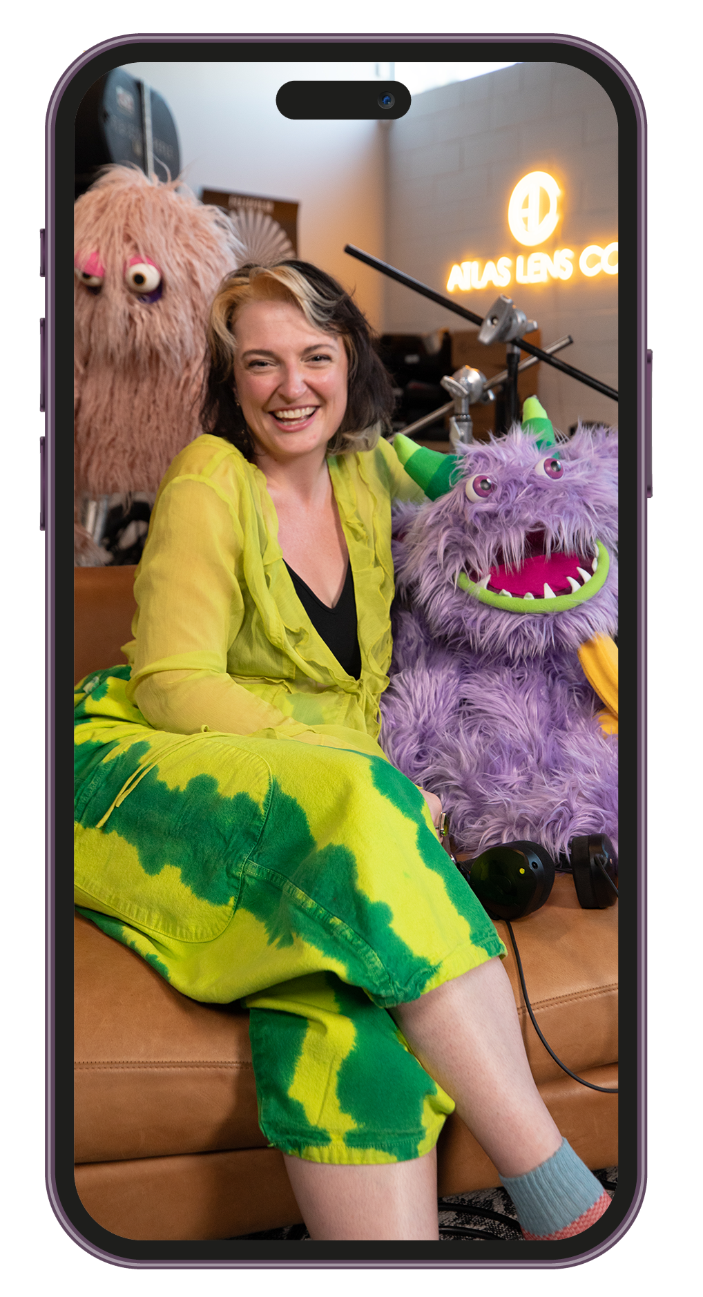 A smiling woman with dark and blonde hair, wearing a yellow and green tie-dye outfit, sitting next to a purple furry monster puppet with green horns and a big mouth, inside a room with a neon sign on the wall.