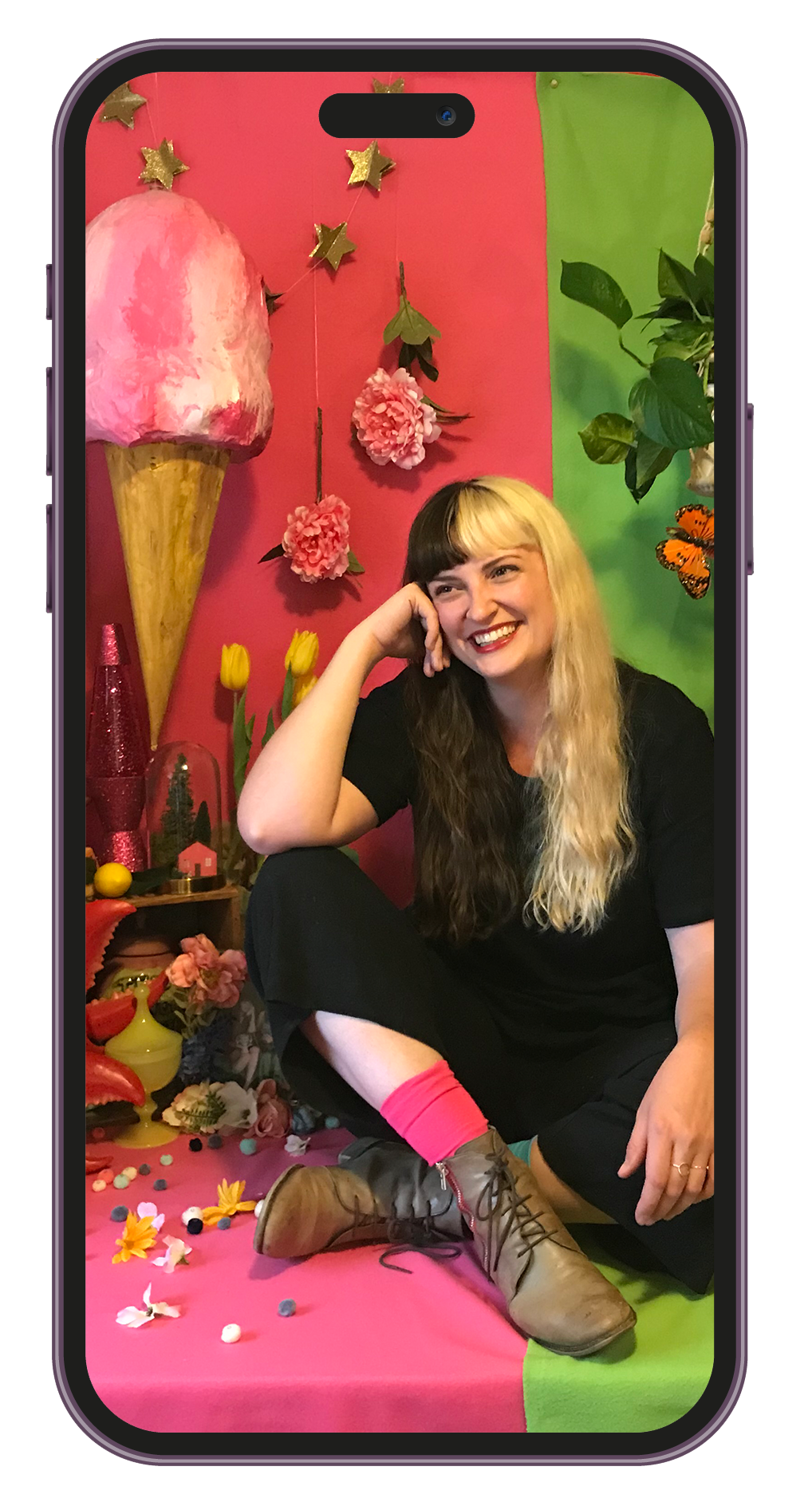 A woman with long, divided hair, blonde on one side and dark on the other, sitting on a pink and green background decorated with flowers, stars, and various colorful objects. She is smiling and dressed in black with pink socks and brown boots.