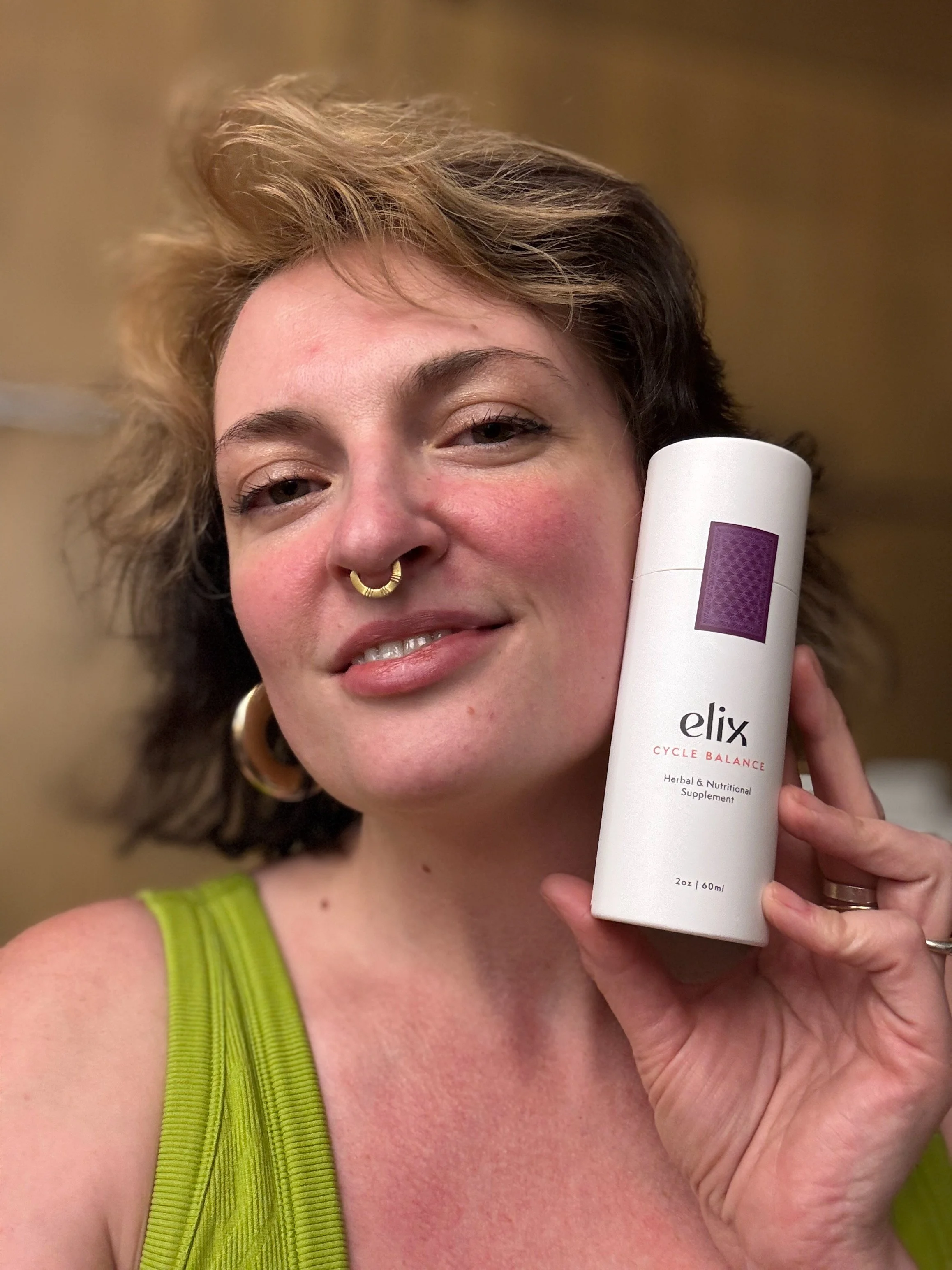 A woman with short, curly hair and a septum piercing holding a white cylindrical bottle labeled 'elix Cycle Balance' close to her face, wearing a lime green tank top.