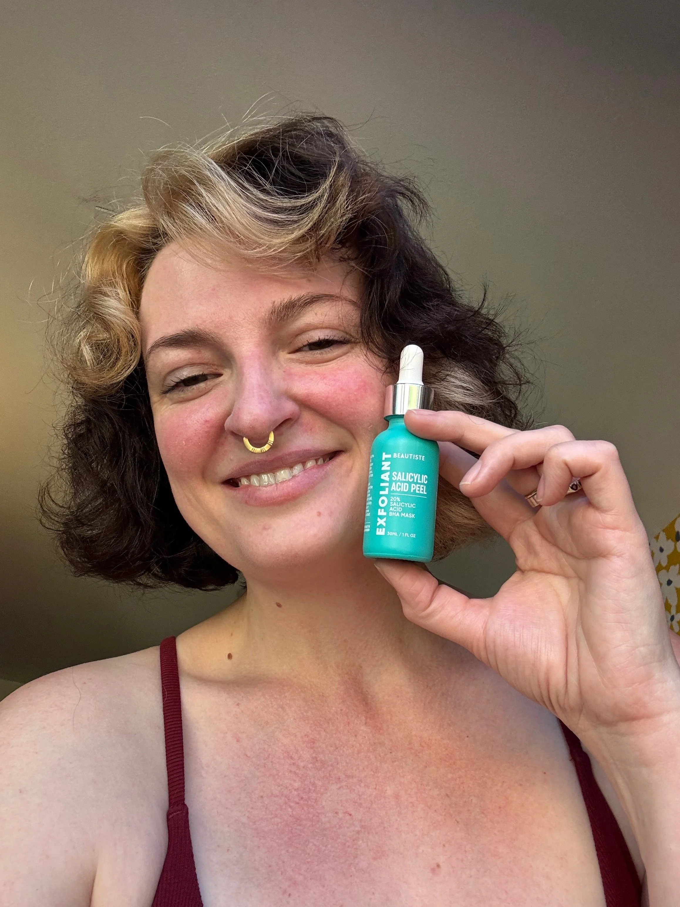 A woman with a mixed dark and blonde curls holding a turquoise bottle of Salicylic Acid Peel, smiling, wearing a red strap top.