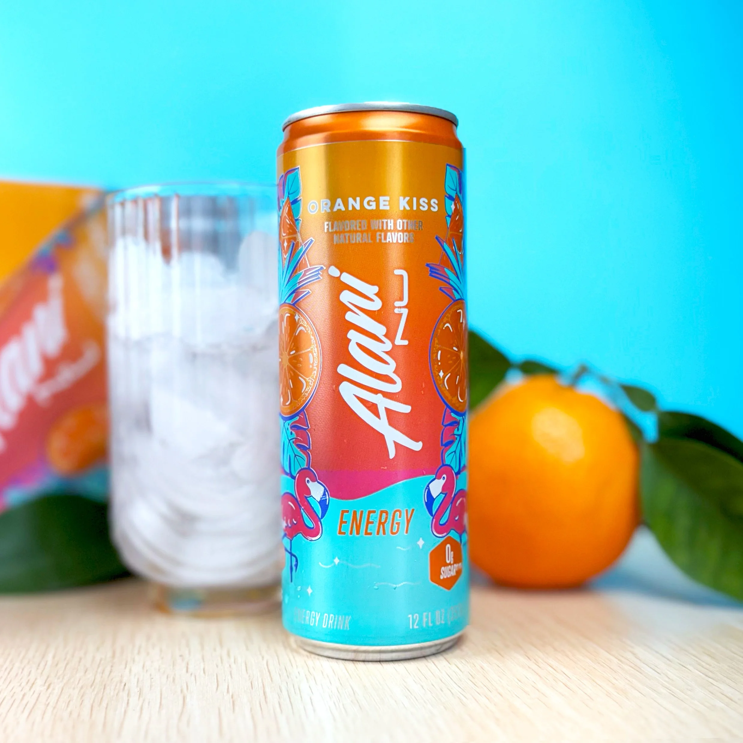 A close-up of an orange energy drink can with blue and white accents, labeled 'Orange Kiss,' on a wooden surface against a light blue background. Part of another similar can and some oranges are also visible in the background.