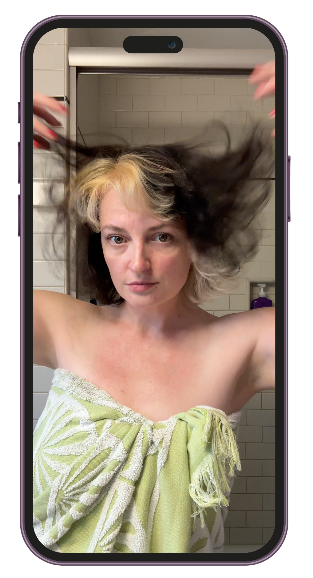 Woman with short, split-color hair, wearing a towel, taking a selfie in a bathroom with beige tiles, holding her hair out to the sides.