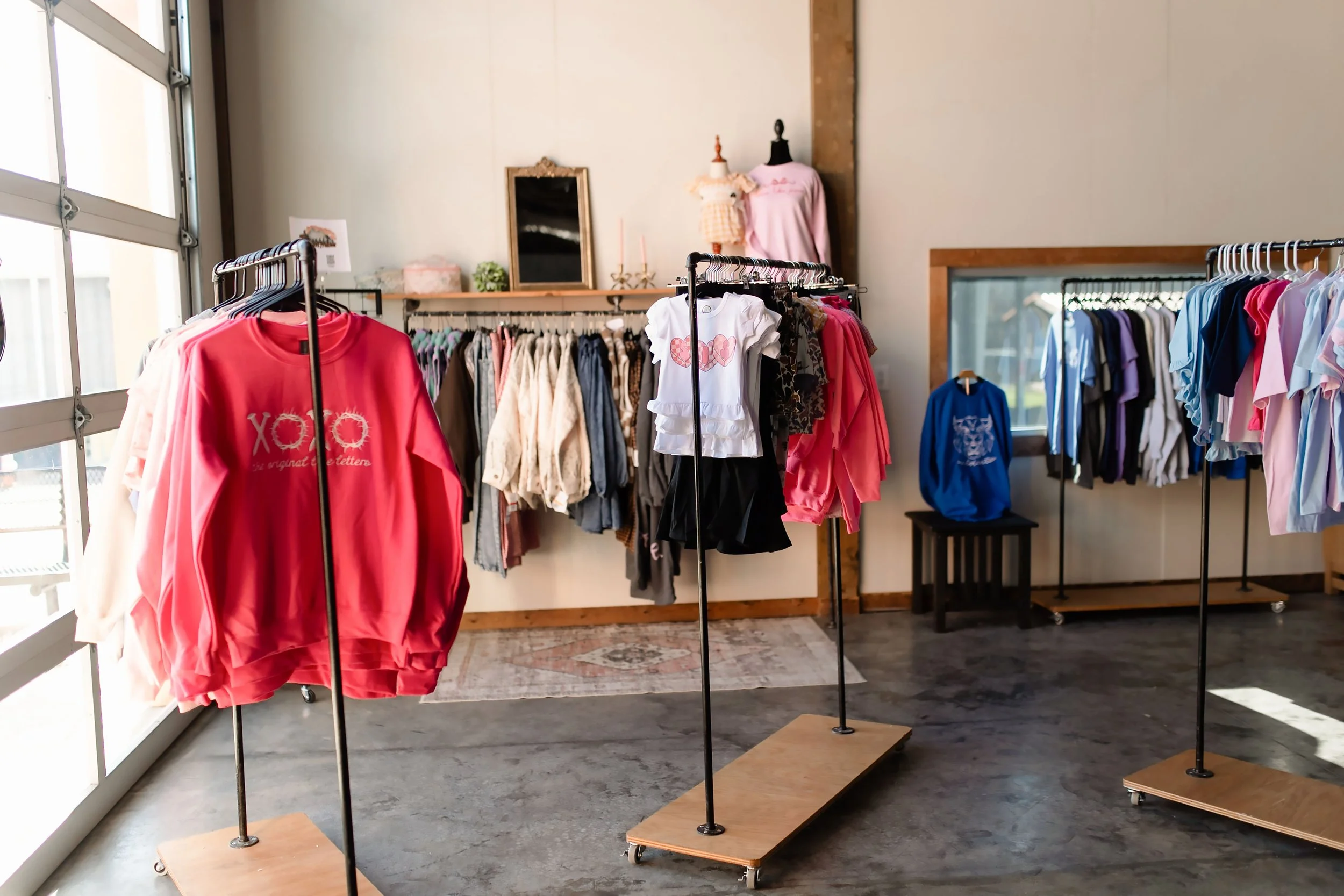 Clothing store with racks of colorful apparel, including sweatshirts, t-shirts, and dresses, inside a well-lit space with large windows and wooden flooring.