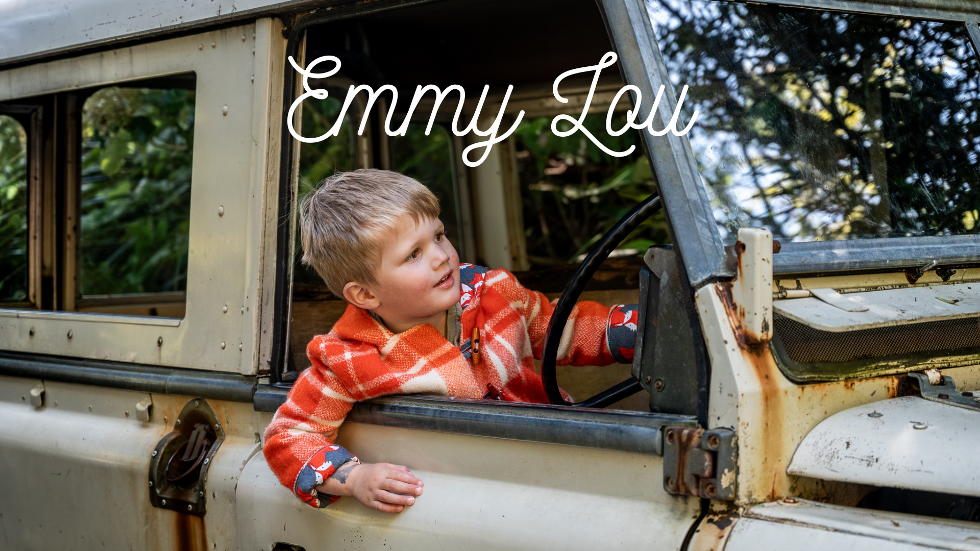 Child in an orange check coat leaning out of a rusty, vintage off-road vehicle with brown and green foliage in the background. The name 'Emmy Lou' is written across the top of the image.