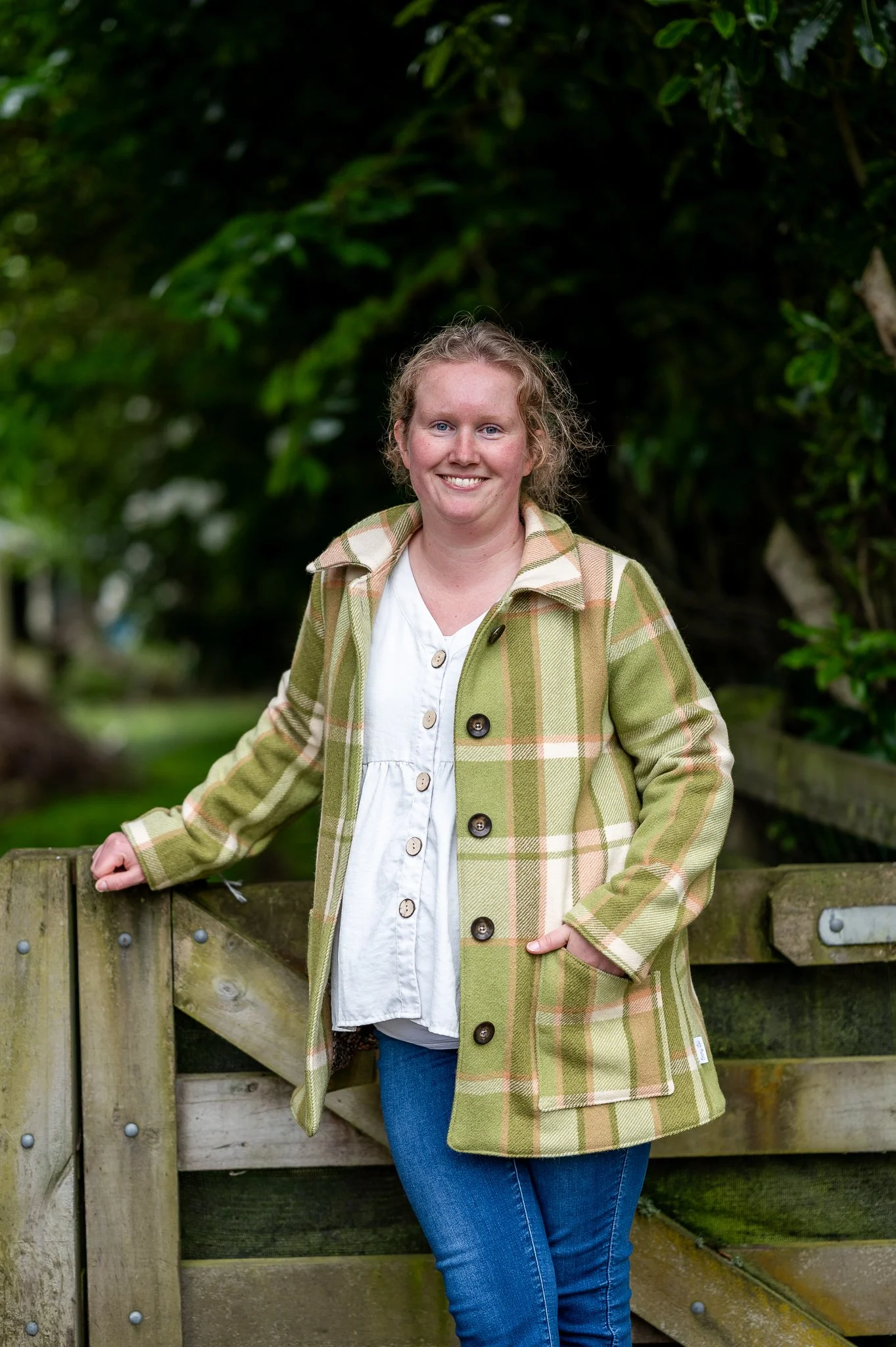 A woman with curly blonde hair wearing a green upcycled wool blanket check coat and white shirt, smiling while standing outdoors near a wooden fence, with trees and greenery in the background.