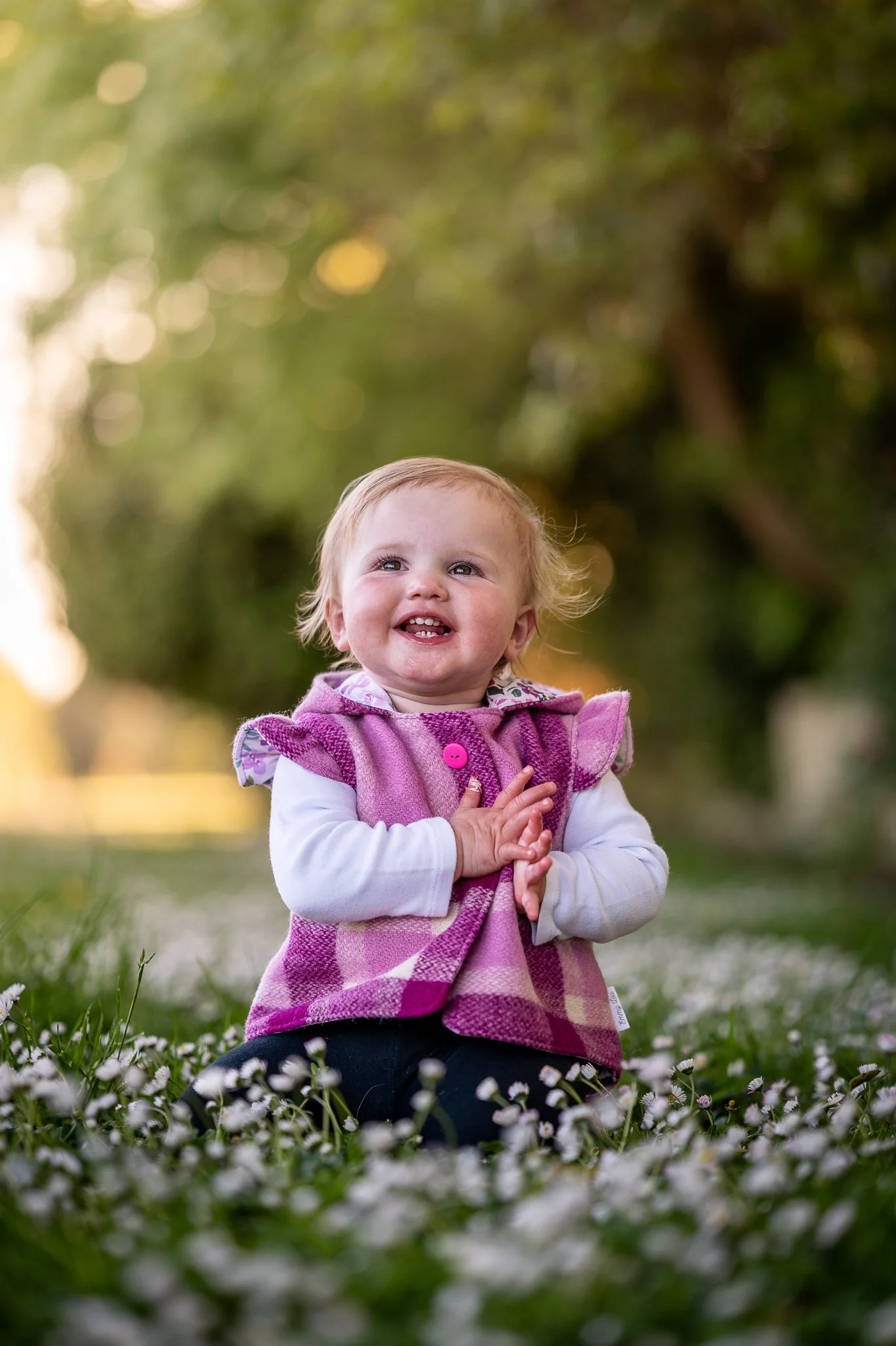 A smiling toddler girl wearing a pink checkered wool blanket vest and white long-sleeve shirt, sitting on grass with small white daisies, outdoors in a park.