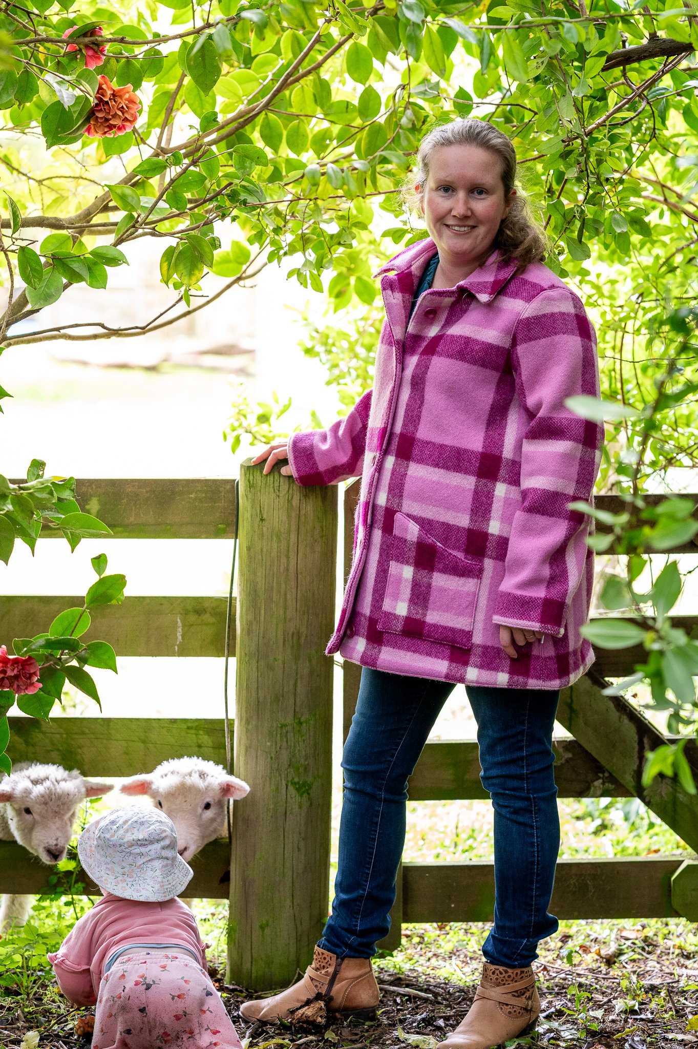 A woman standing outdoors next to a wooden fence, smiling, wearing a pink check wool coat, with little lambs near her, a child in a pink outfit and hat looking at the lambs, surrounded by green foliage.