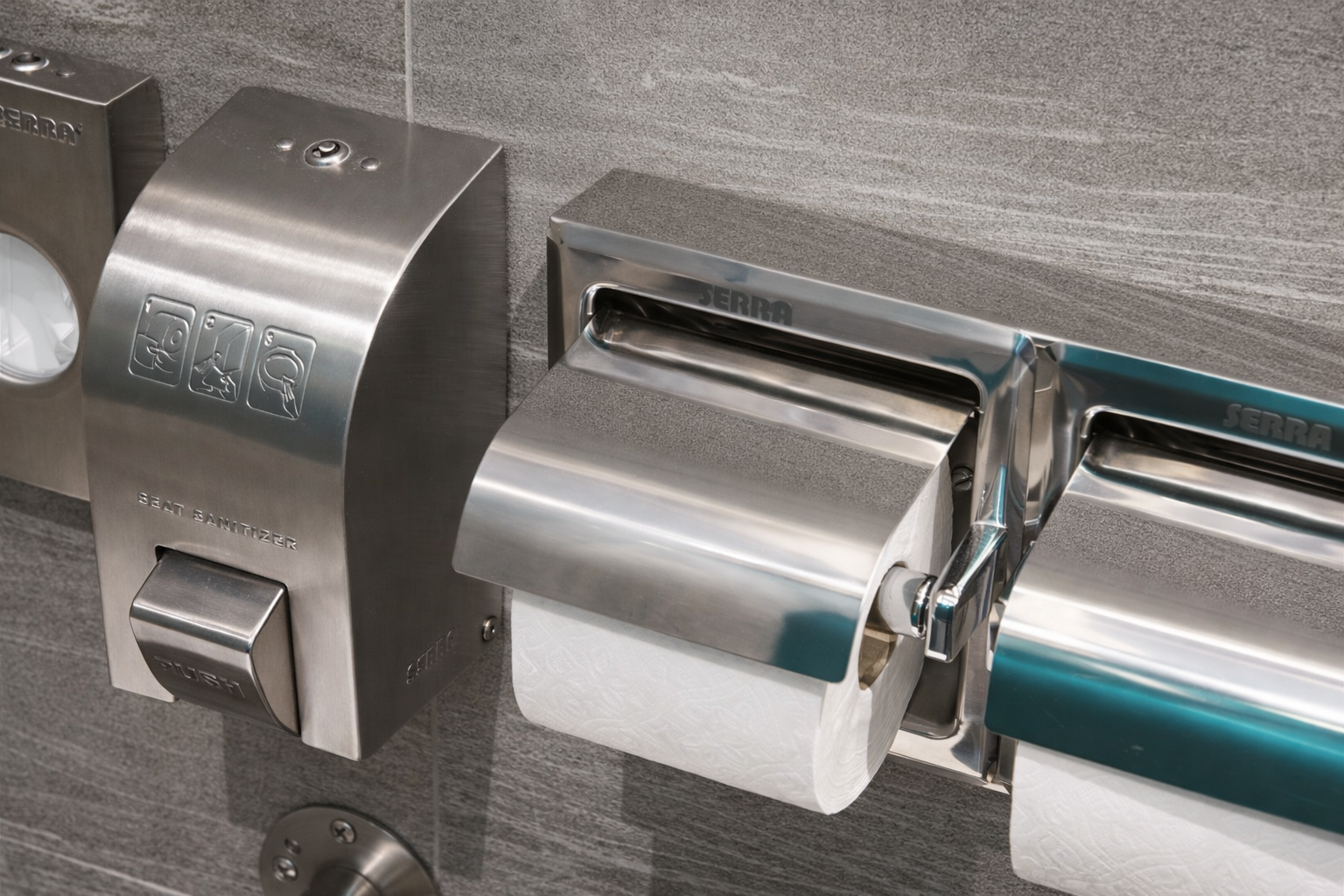 Stainless steel toilet paper dispenser and a manual seat sanitizer on a gray tiled wall.