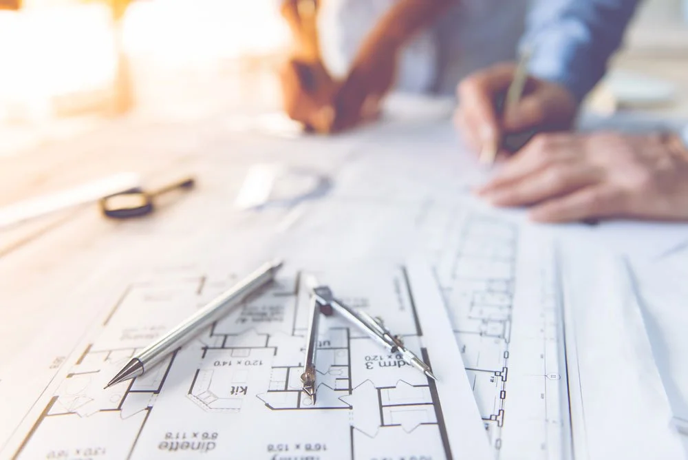 Architectural blueprints with pens and drafting tools on a table, person in the background sketching.