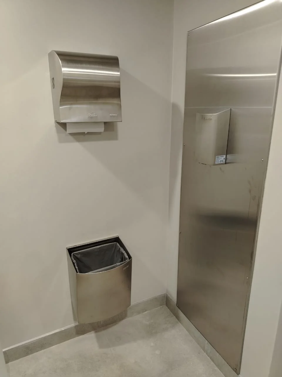 Stainless steel paper towel dispenser, trash bin in a restroom or hallway corner.