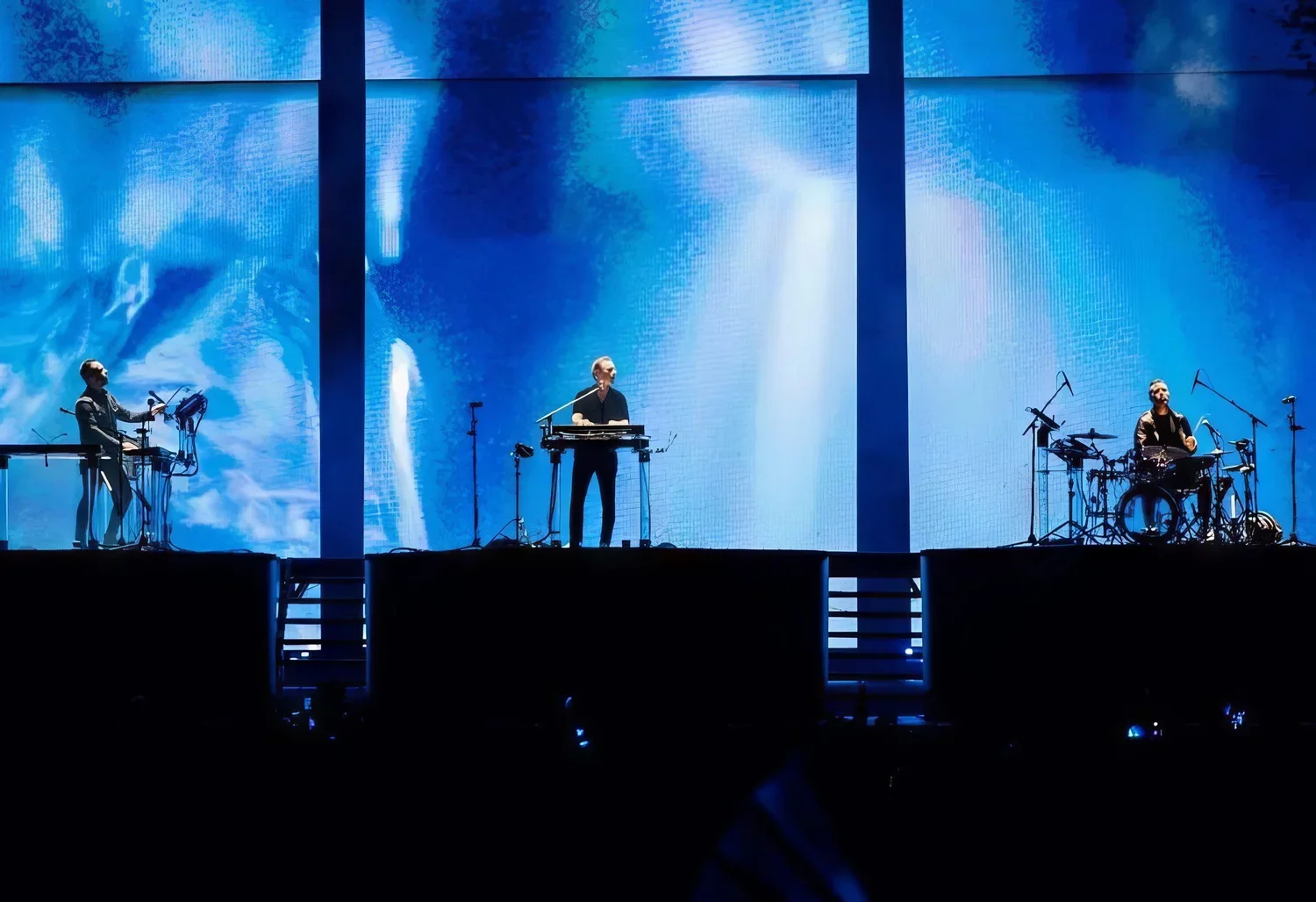 Three musicians performing on a stage with a digital blue background. One on the left is adjusting electronic equipment, the middle is standing behind a keyboard, and the right is sitting at a drum set. They are illuminated by stage lighting.