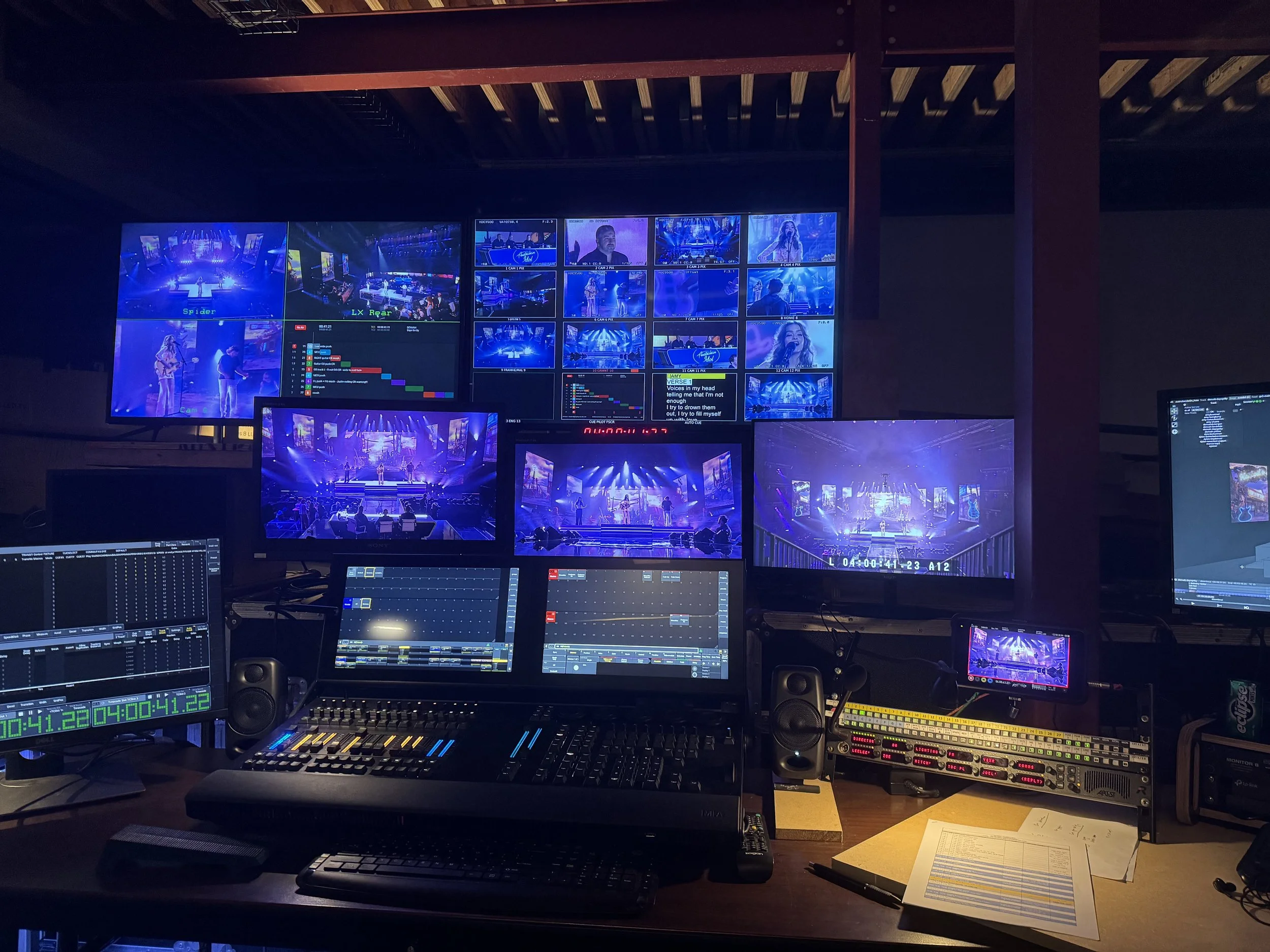 A control room with multiple monitors displaying a concert or performance with stage and performers, along with technical equipment and control panels.