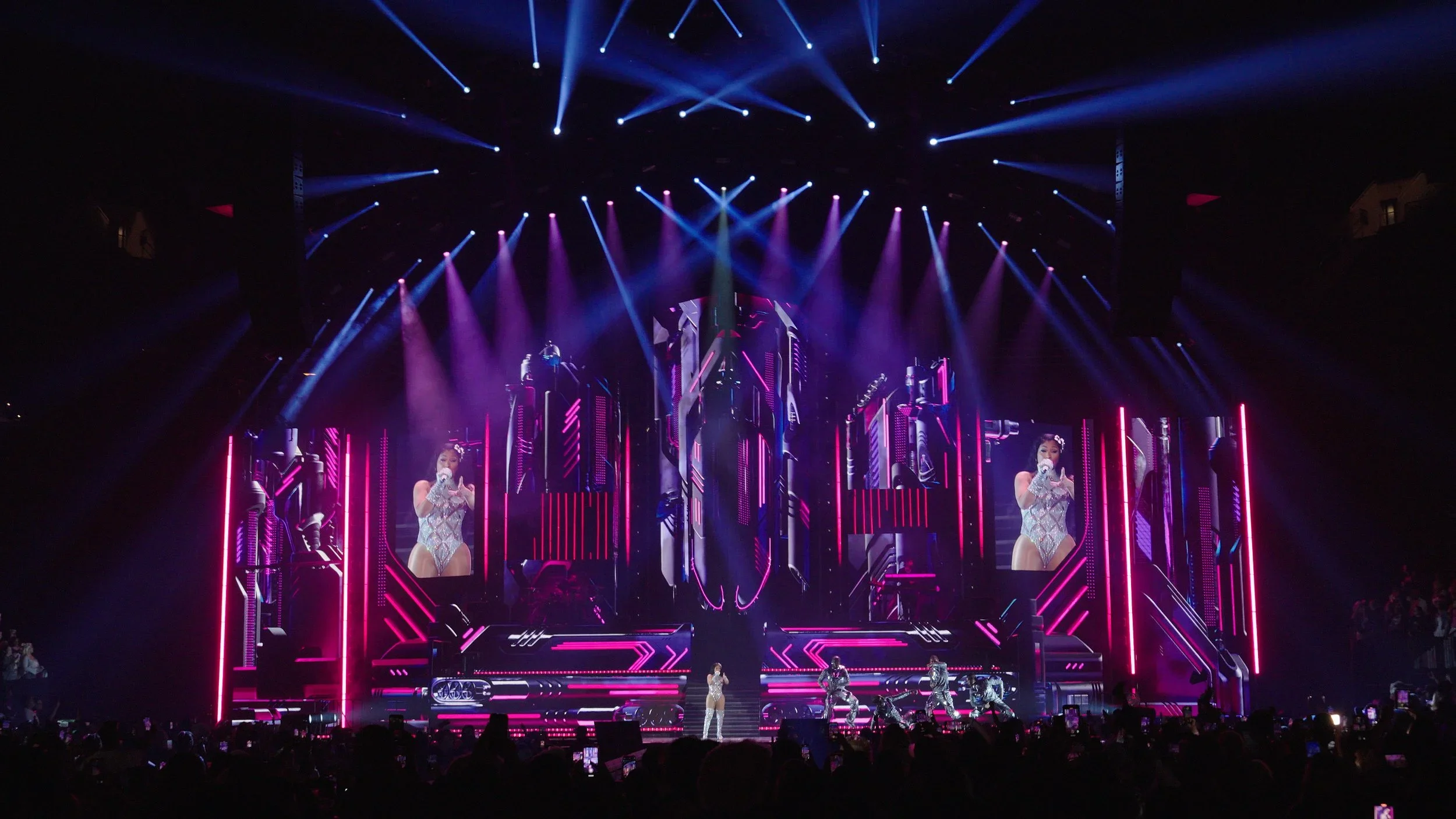 A concert stage with colorful neon lights and multiple large screens displaying performers, including a singer in a silver outfit, with an audience in front.