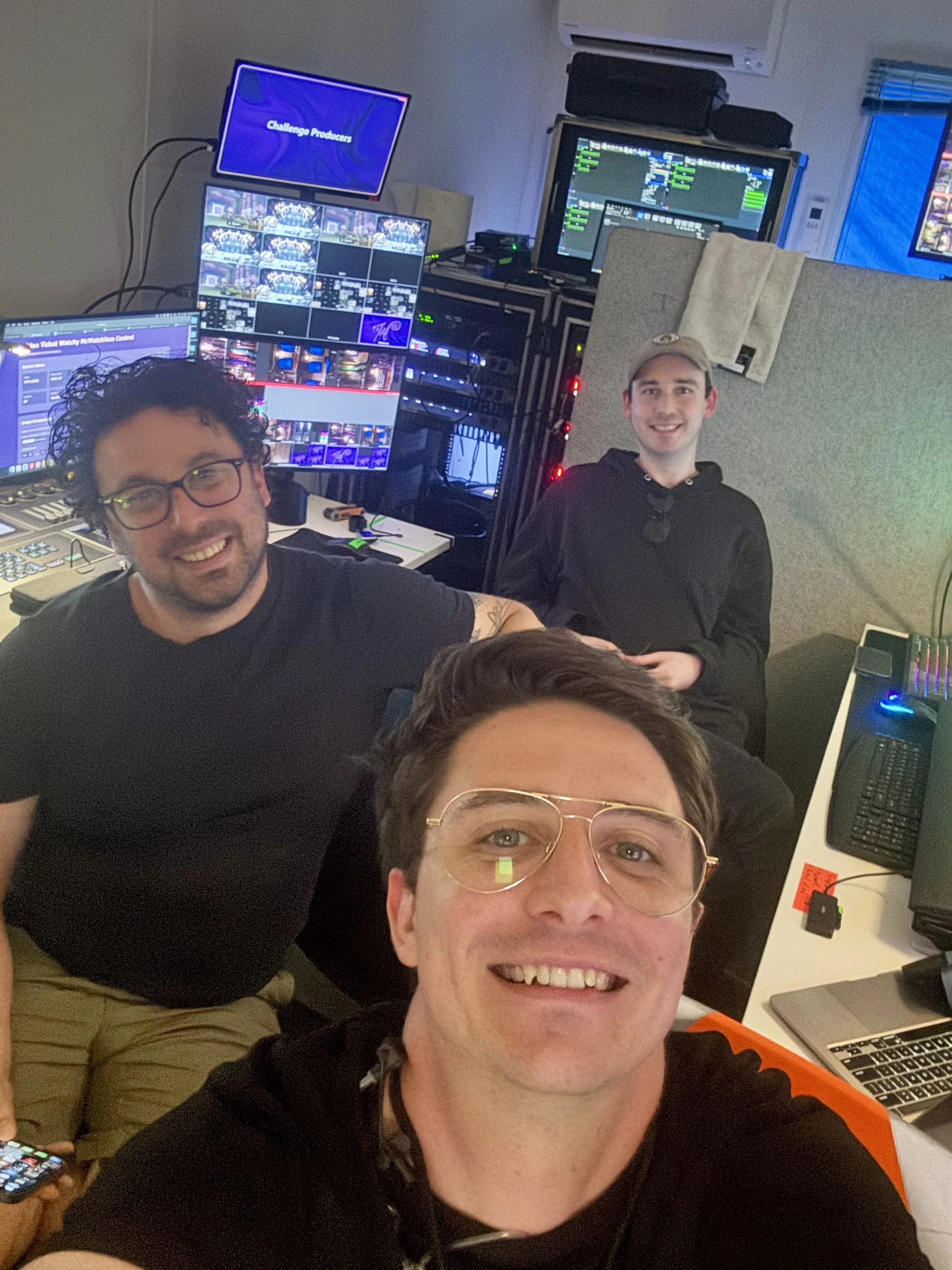 Group of four people in a room with multiple computer monitors, audio equipment, and a TV screen displaying 'Challenge Producers.' They are smiling, with one person taking a selfie. The room appears to be a control or broadcast setup.