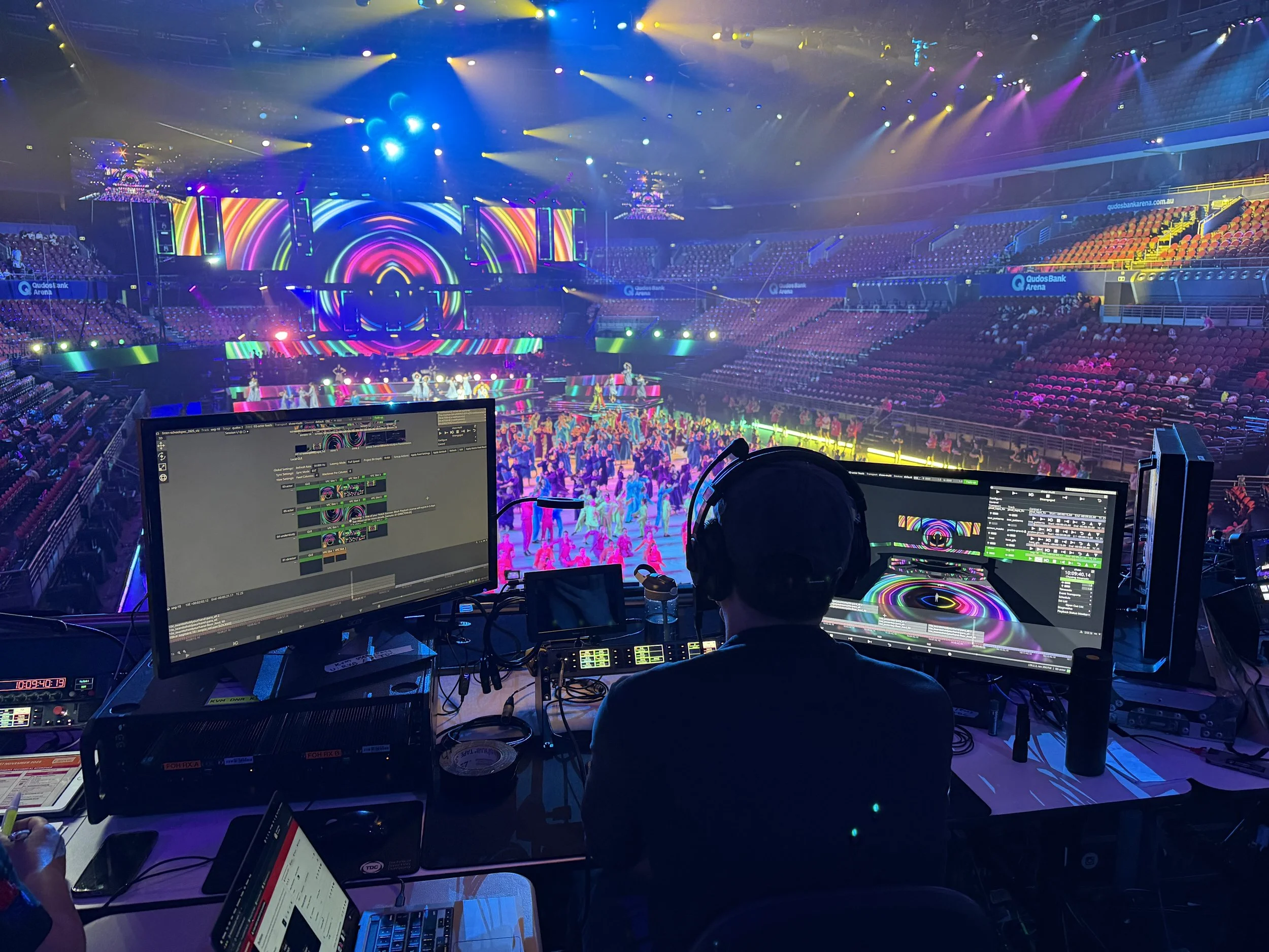 A stage with colorful lighting and digital screens in an arena, with performers and audience seated around the stage. The photo is taken from the control booth with multiple monitors showing event controls.