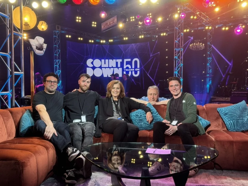 Group of five people sitting on a sofa at a colorful event stage with a sign reading 'Count 50 Years on' and various stage lights and decorations in the background.