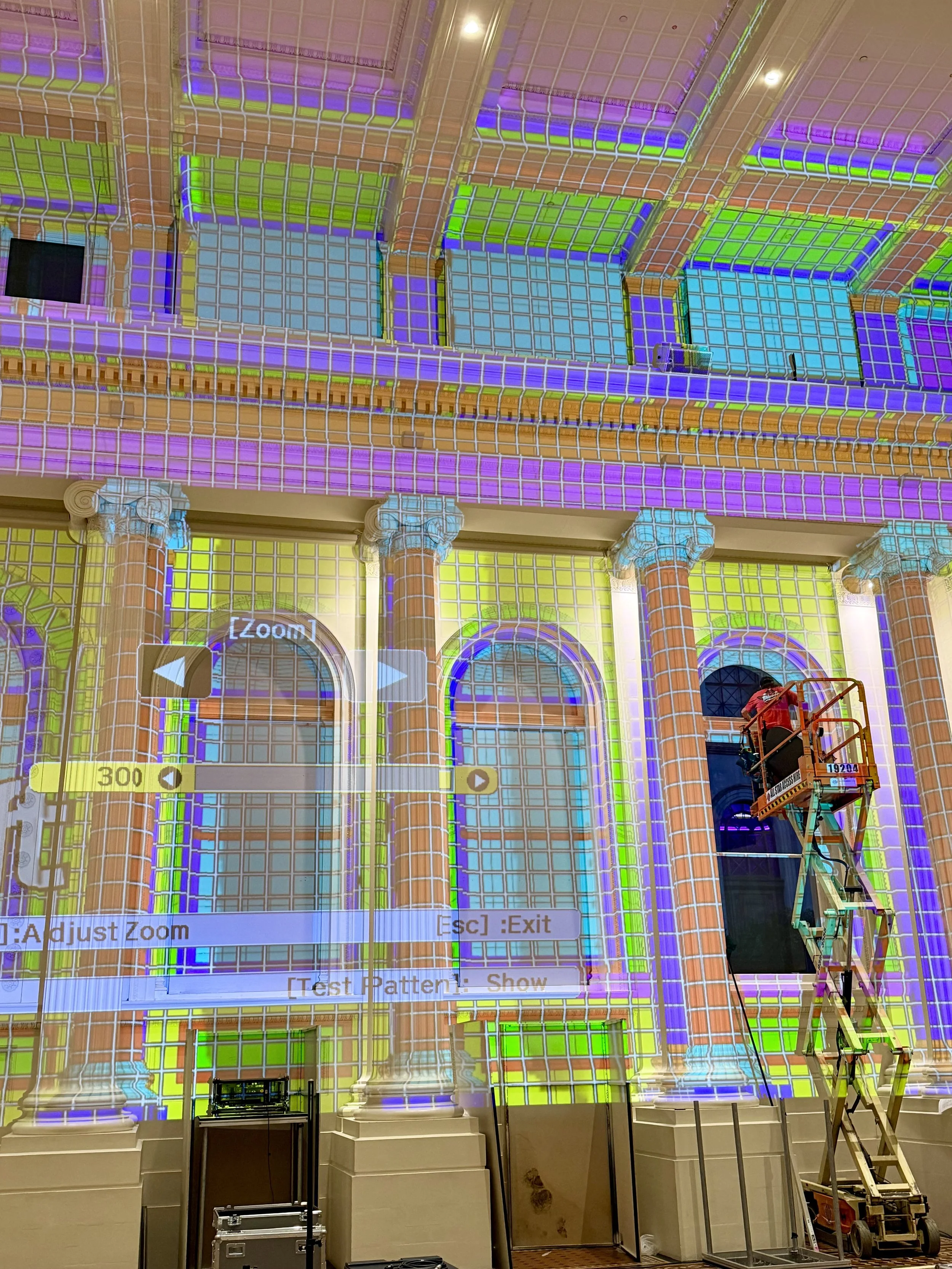 Projection mapping on a building's facade showing colorful patterns and architectural details, with a worker on a lift.