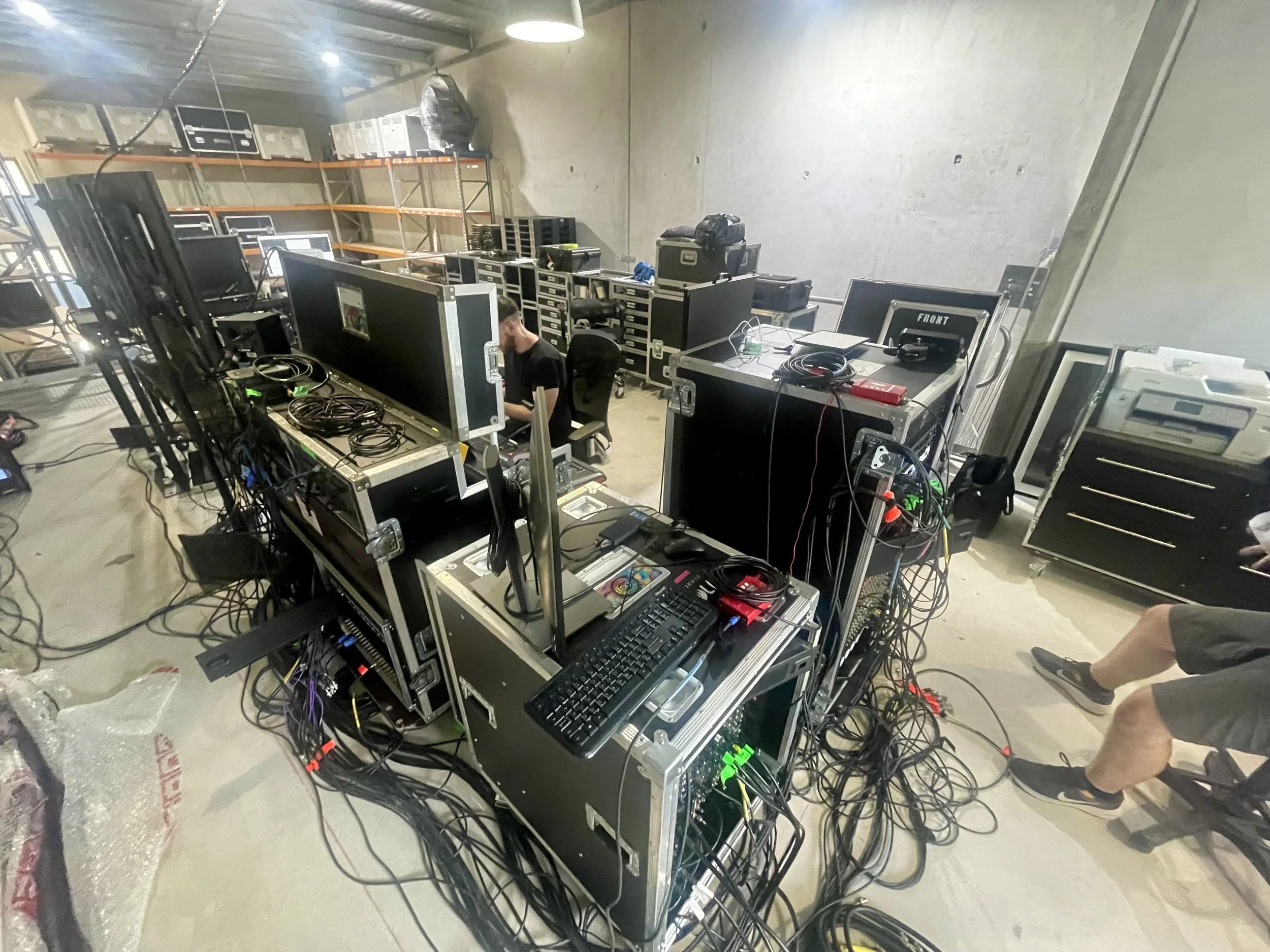A person working at a desk with multiple monitors, routers, and electronic equipment, surrounded by cables in an unfinished industrial space.