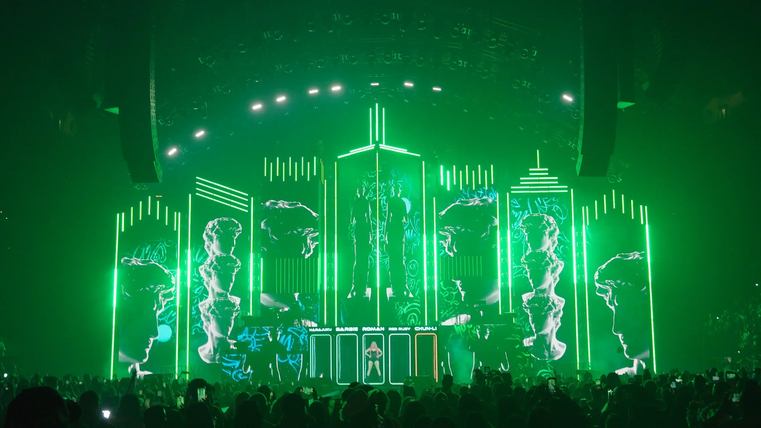 A concert stage with vivid green lighting, featuring stylized images of classical sculptures' heads and a central figure, with a backdrop displaying names of various artists and performers.