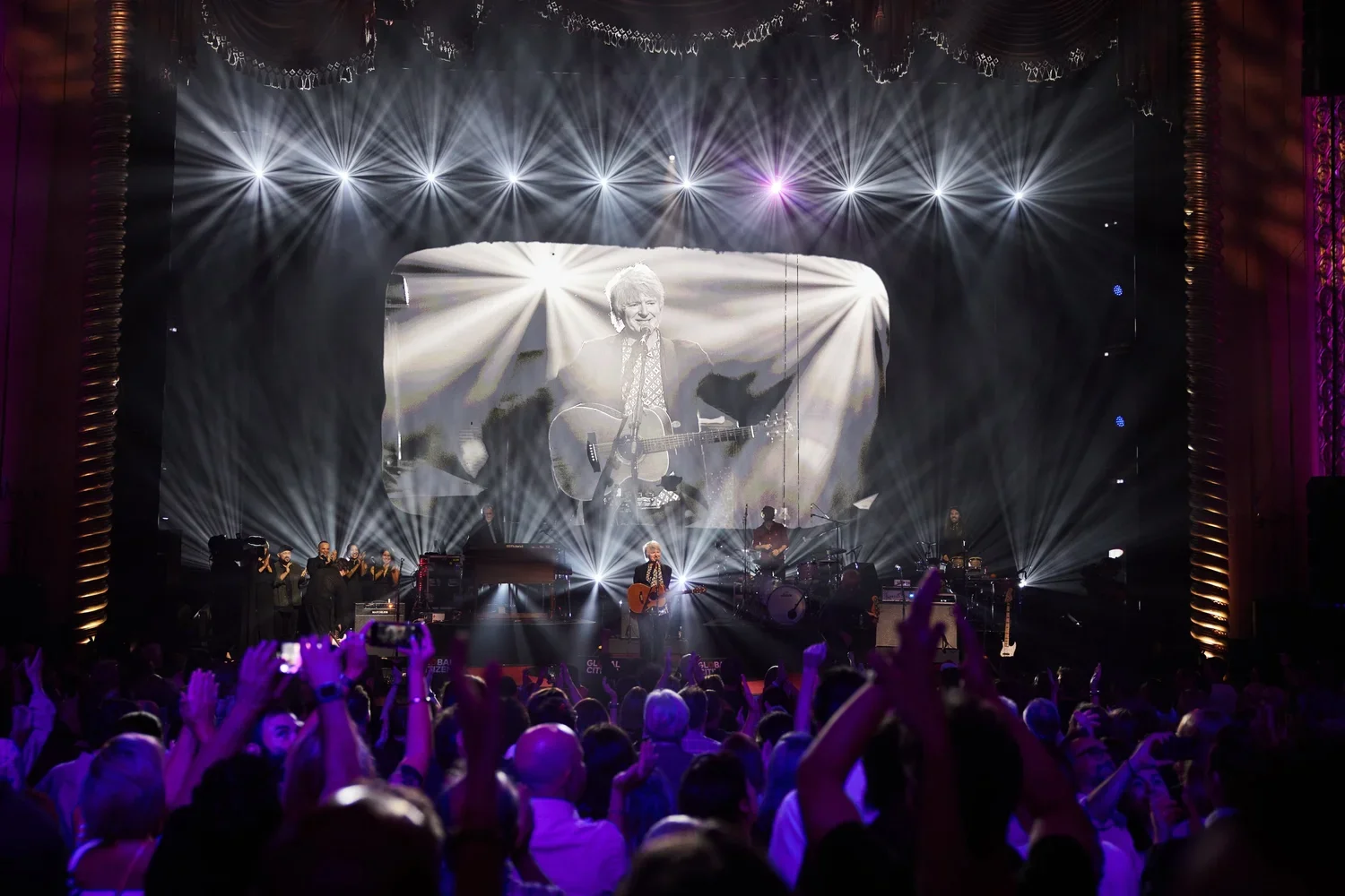 Concert scene with a performer playing guitar on stage, large screen displaying a black-and-white image of the same performer, crowd with raised hands, stage lighting, and backup musicians.