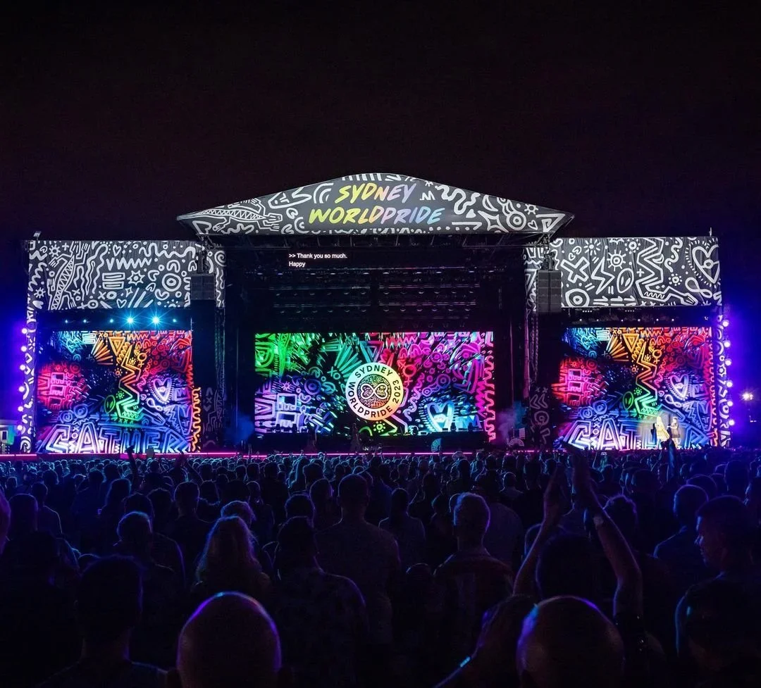 Nighttime scene of an outdoor concert stage decorated with colorful, neon-style artwork and text, with a crowd of people watching in the foreground.