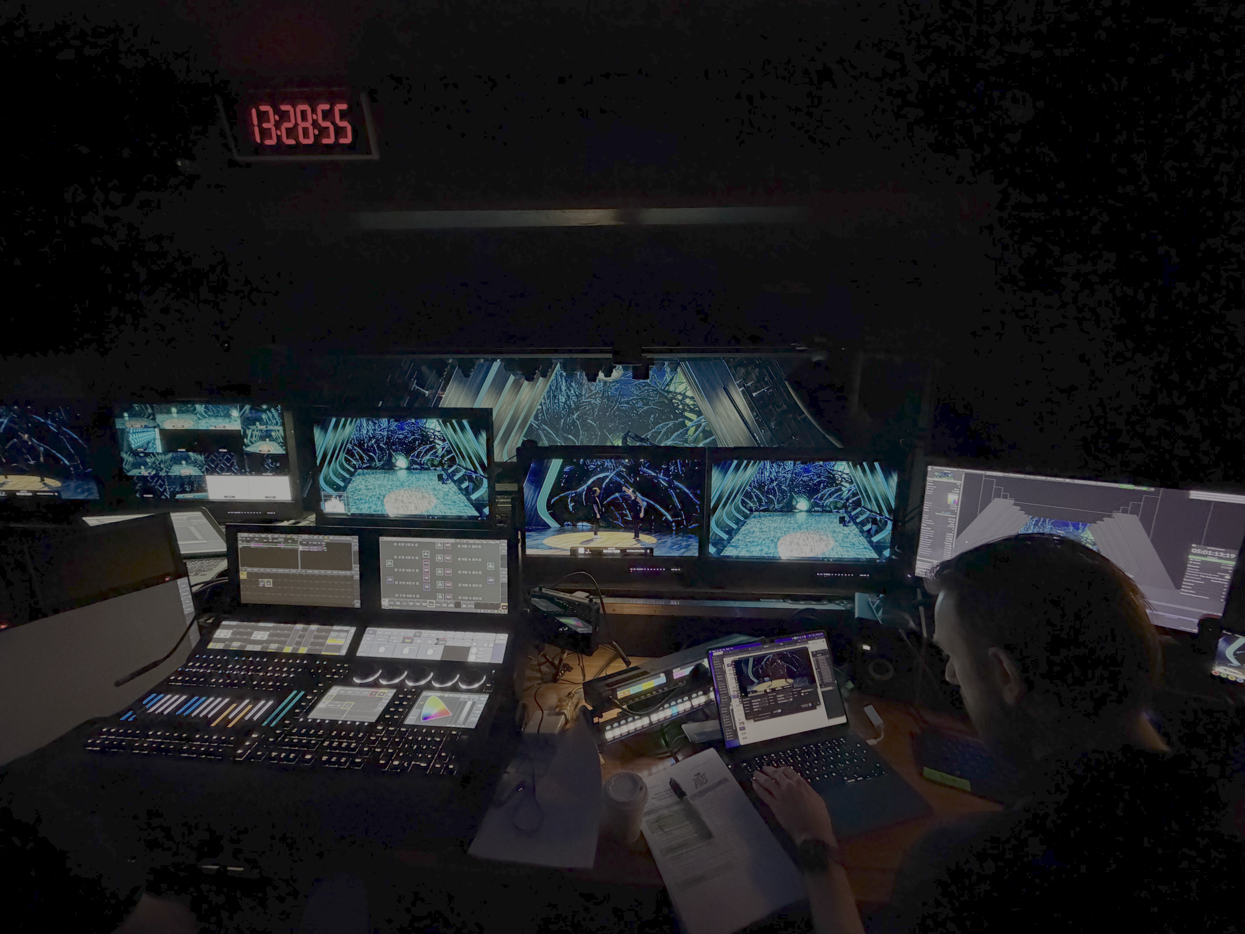Dark room with multiple computer monitors displaying digital designs and visual effects, and a person working on a computer.