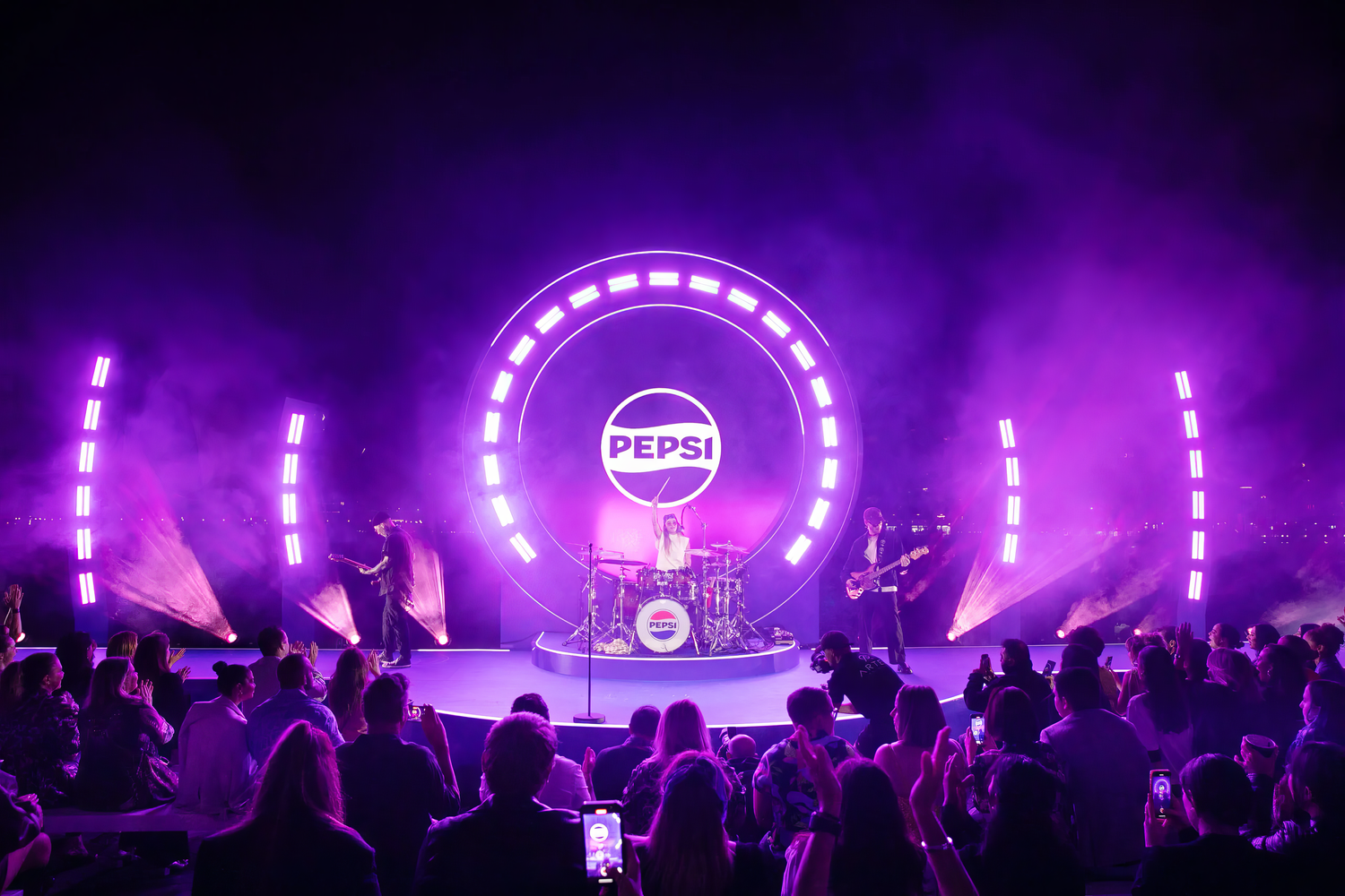 A live music concert with a three-member band performing on a stage illuminated with purple lights. The backdrop features a large circle with the Pepsi logo inside. The audience is watching and some are taking photos.