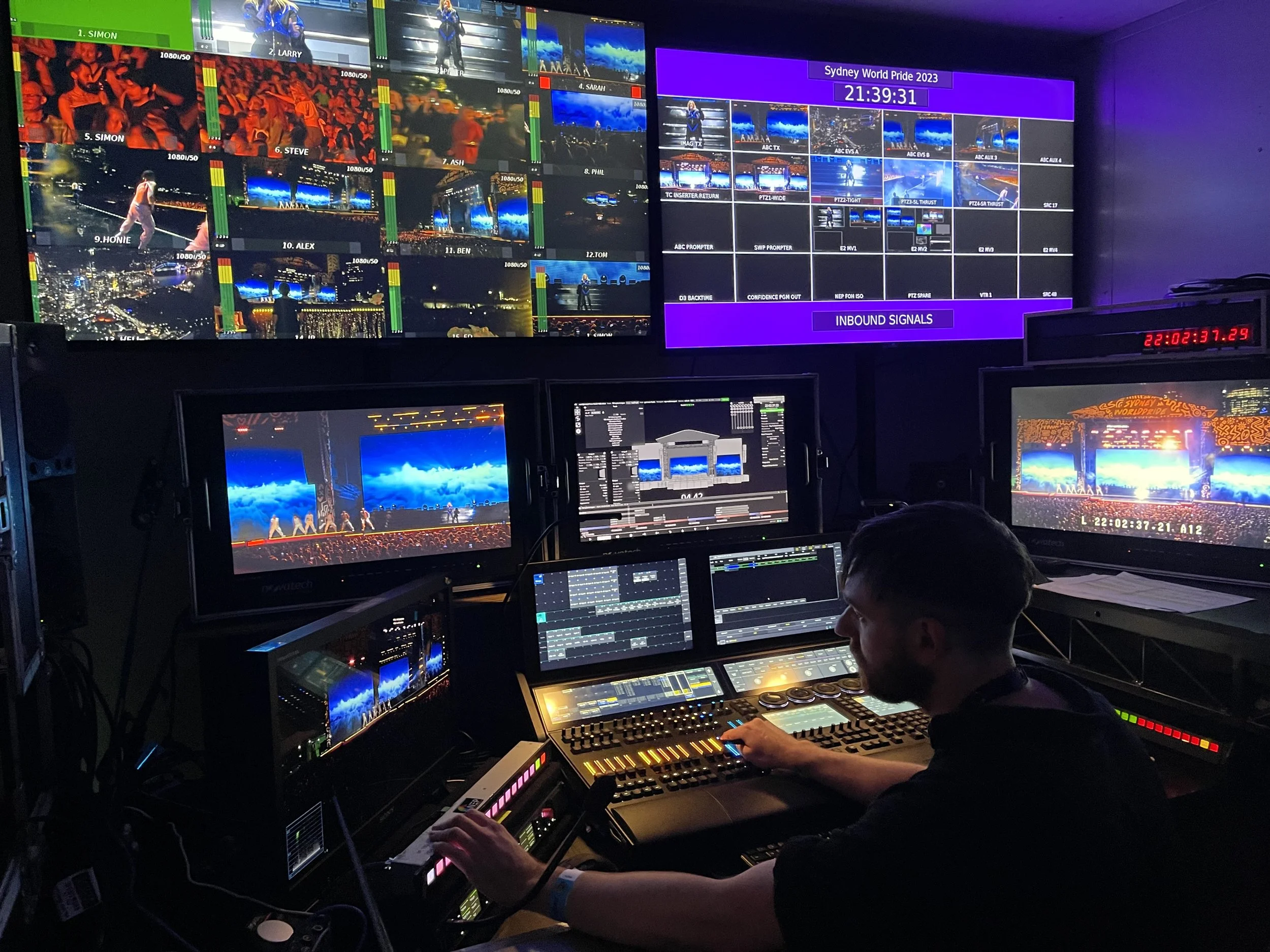 Video control room with multiple monitors displaying video feeds, with a person operating a control panel. The screens show scenes from a live event, labeled 'Sydney World Pride 2023', and the room has vibrant lighting.