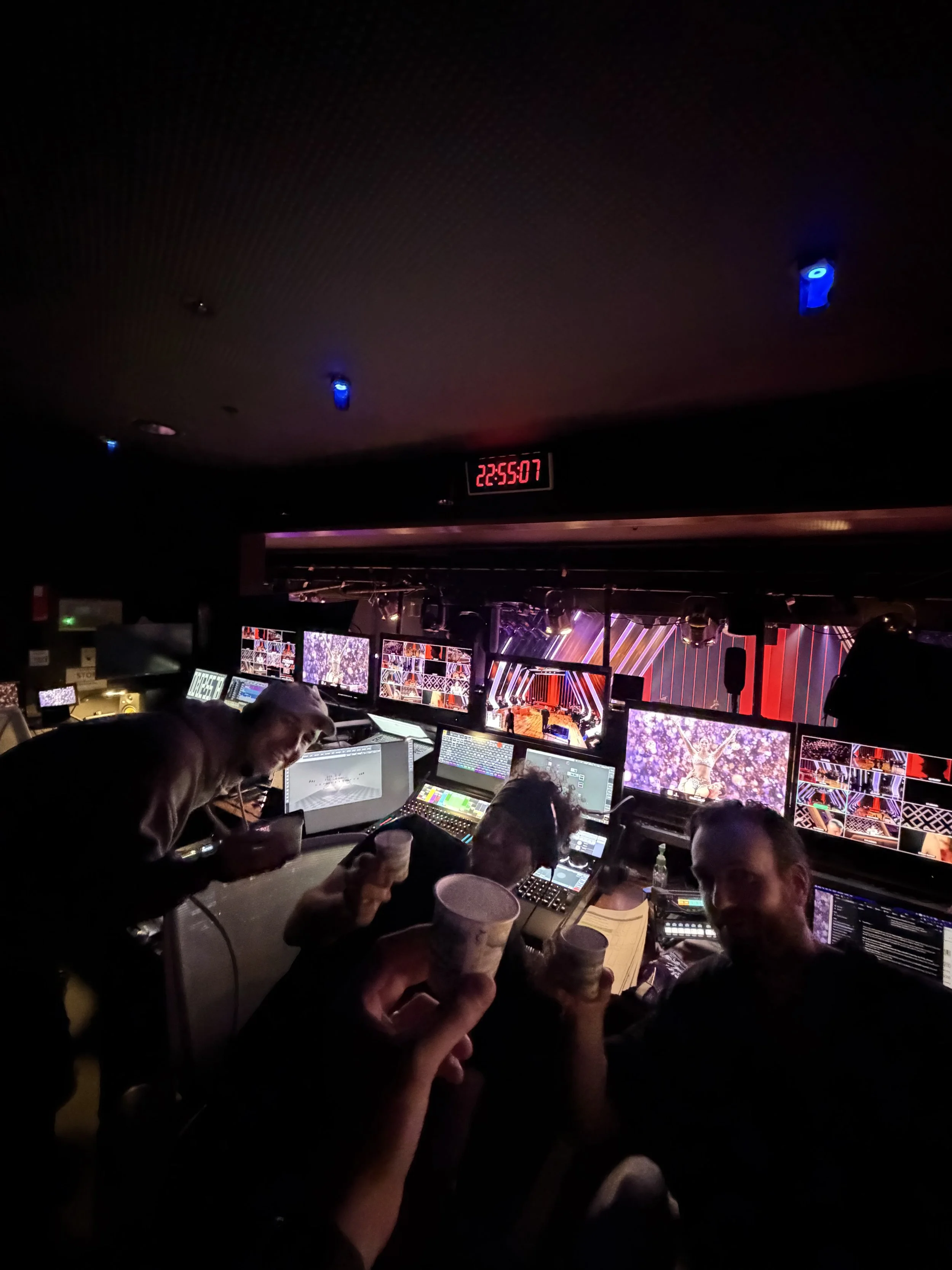 People working at a control room with multiple monitors, a digital clock displaying 22:55:07, and a stage with colorful lighting in the background, possibly during a live event or concert.