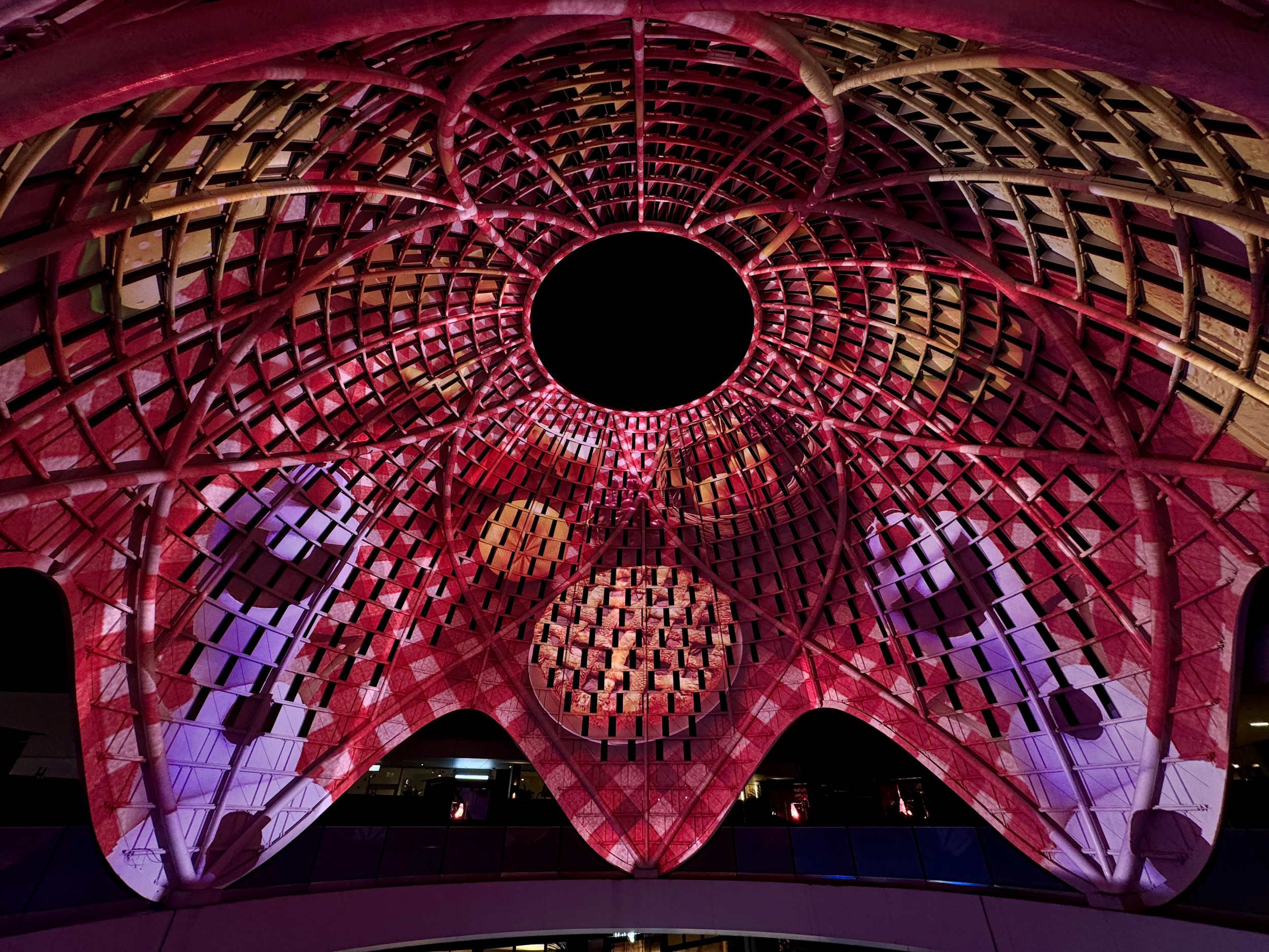 The image shows the interior of a colorful, modern architectural structure with a circular, grid-like pattern on the ceiling, illuminated with purple and pink lighting.