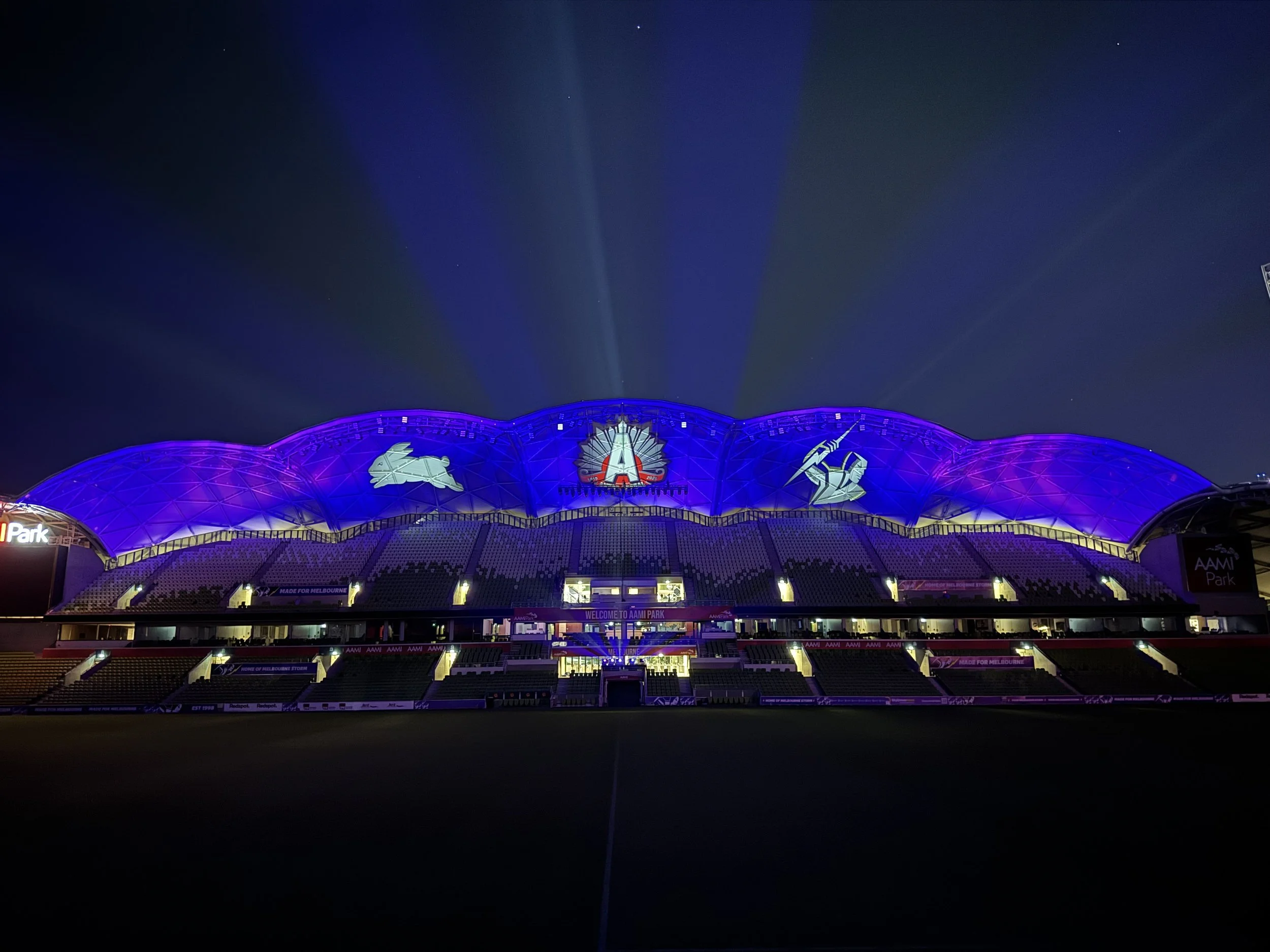 Night view of a stadium with a purple-lit dome and seating area, with the Melbourne Cricket Ground logo illuminated at the center of the dome.