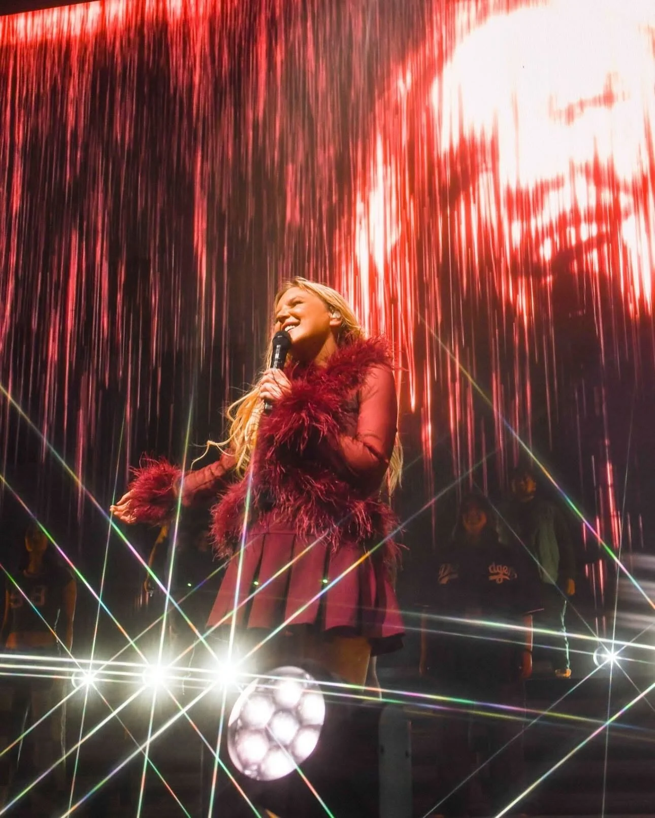 A woman with long blond hair singing into a microphone on stage. She is wearing a red dress with fur trim. Behind her is a red curtain and colorful stage lights.