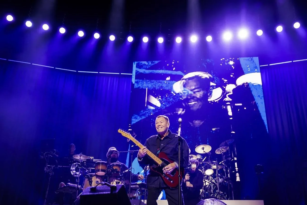 A musician on stage playing an electric guitar, smiling, with a band and drums behind him, and a large screen displaying a close-up of his face in the background, illuminated by purple and white stage lights.