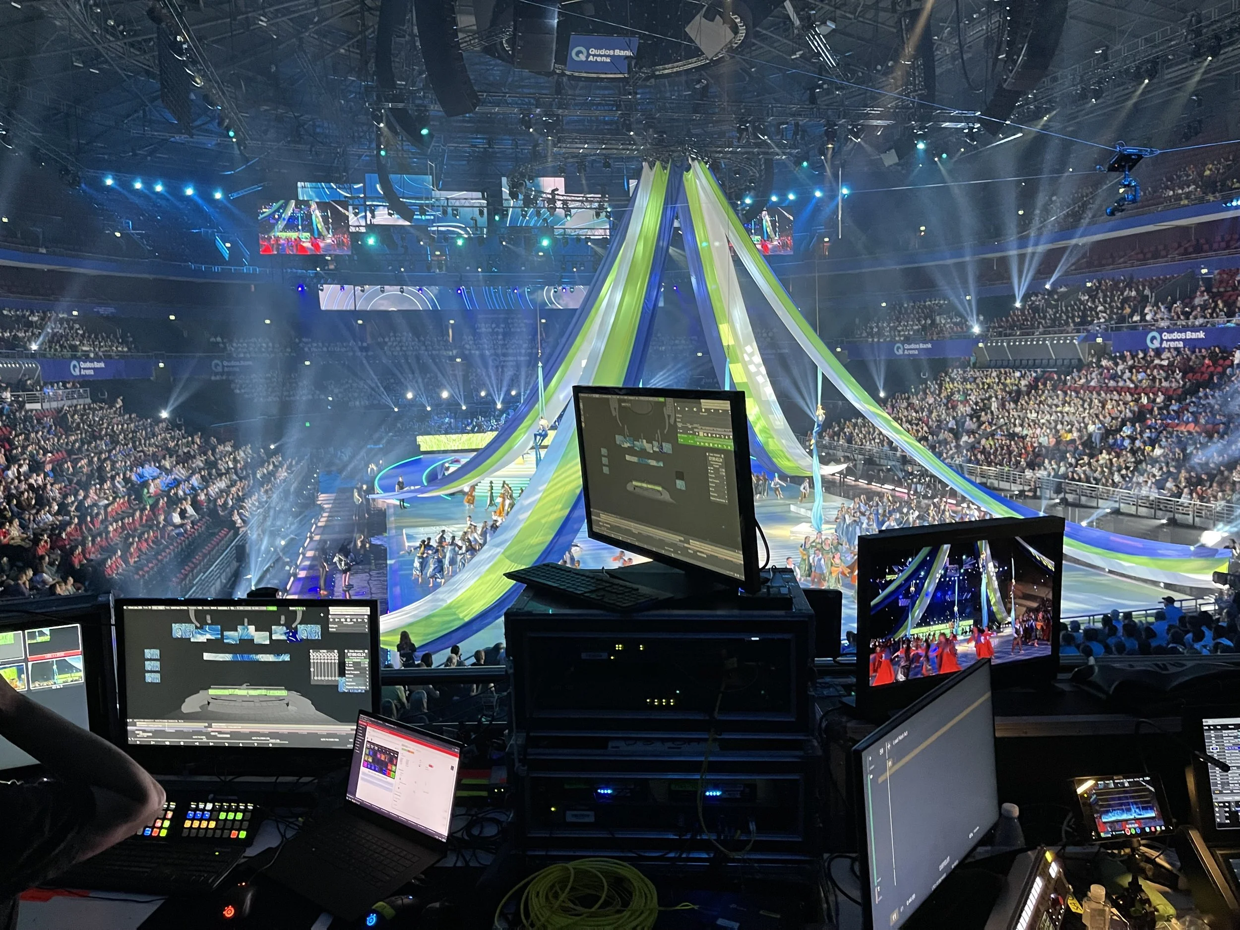 Photo of an indoor event arena with a large audience, stage, and lighting setup. The image shows multiple screens, lighting rigs, and a control panel.