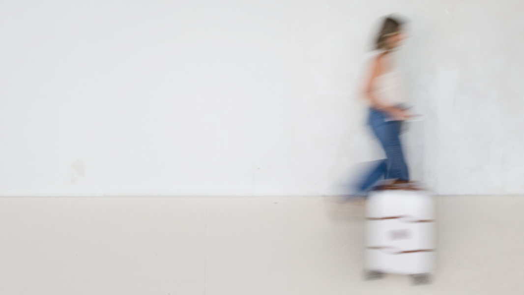 A woman walking with a suitcase in motion, creating a blurred effect, against a plain wall.