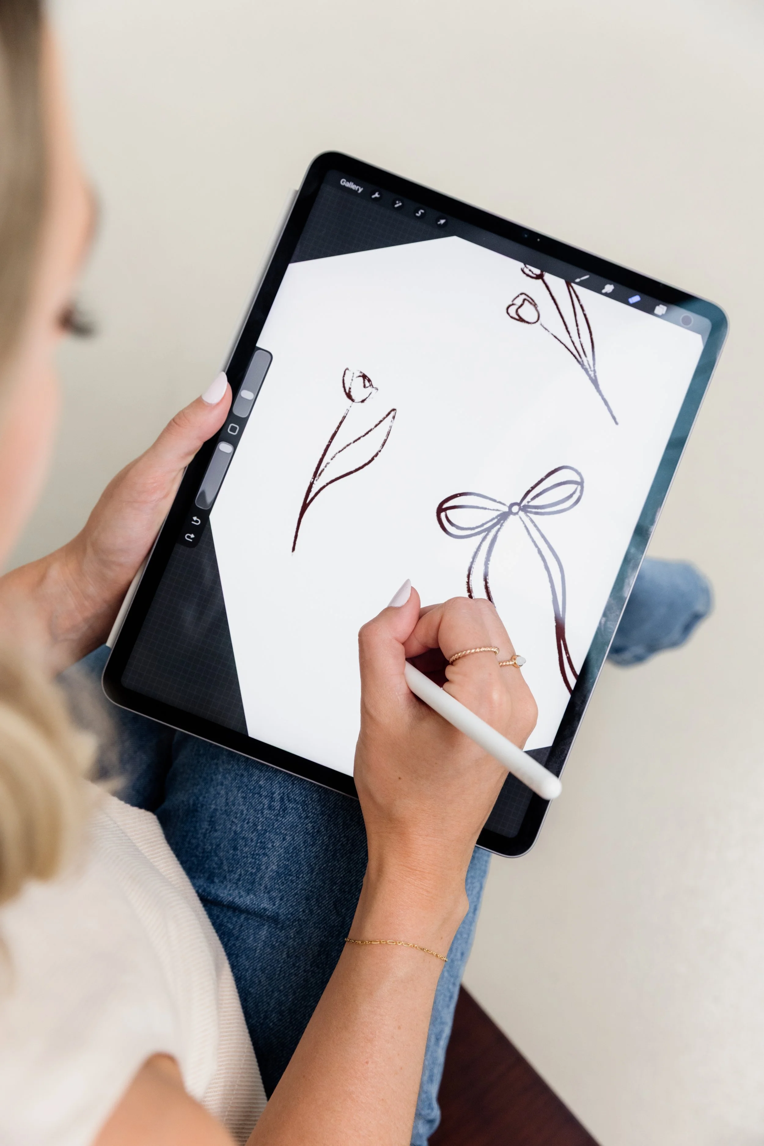 A woman is sitting down, using a tablet with a stylus. She is drawing sketches of flowers and a bow on the tablet screen.