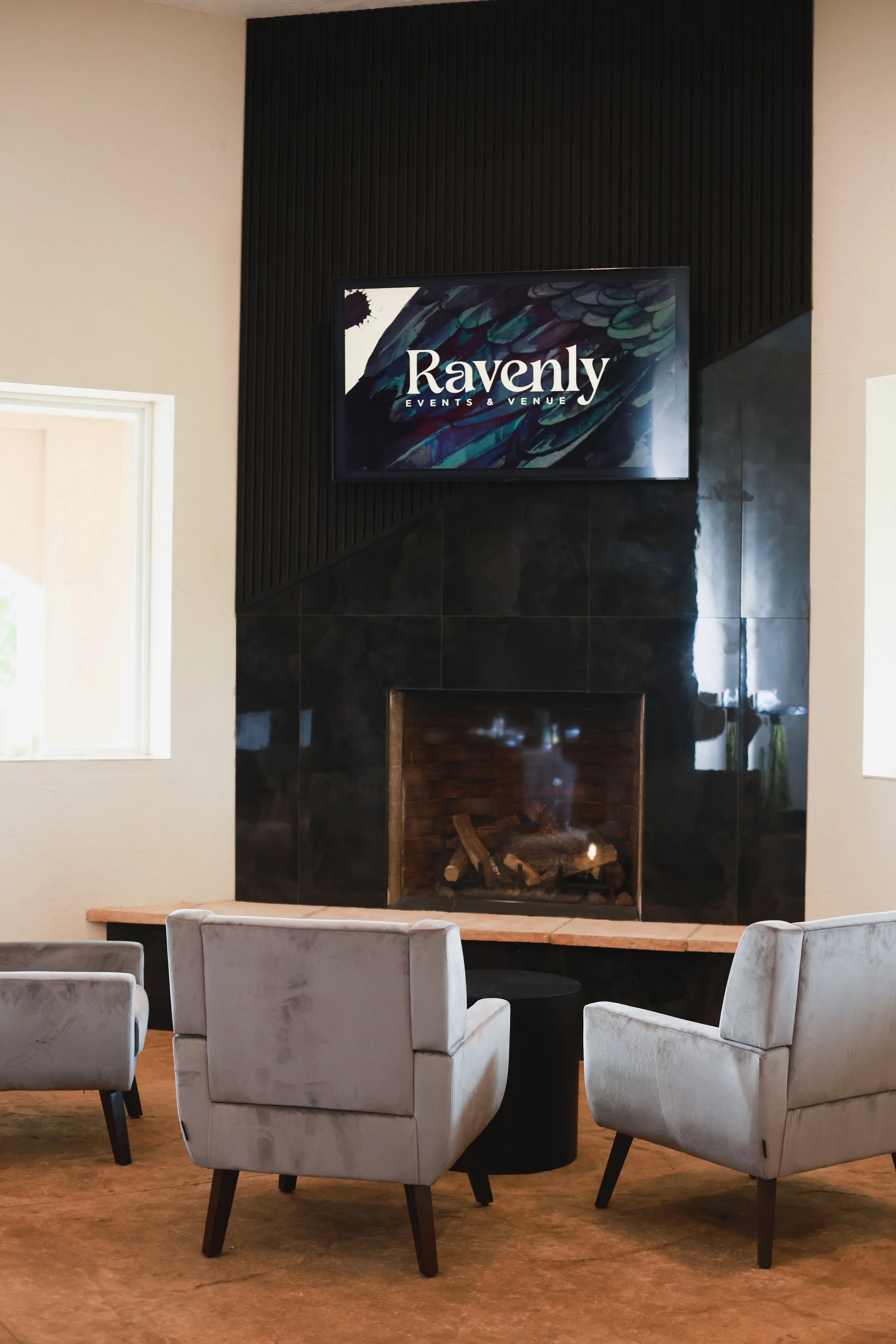A living room with a fireplace, a flat-screen TV above it displaying the text "Ravenly Events & Venue," and four white upholstered chairs with dark wooden legs arranged around a black round table on a wood floor.