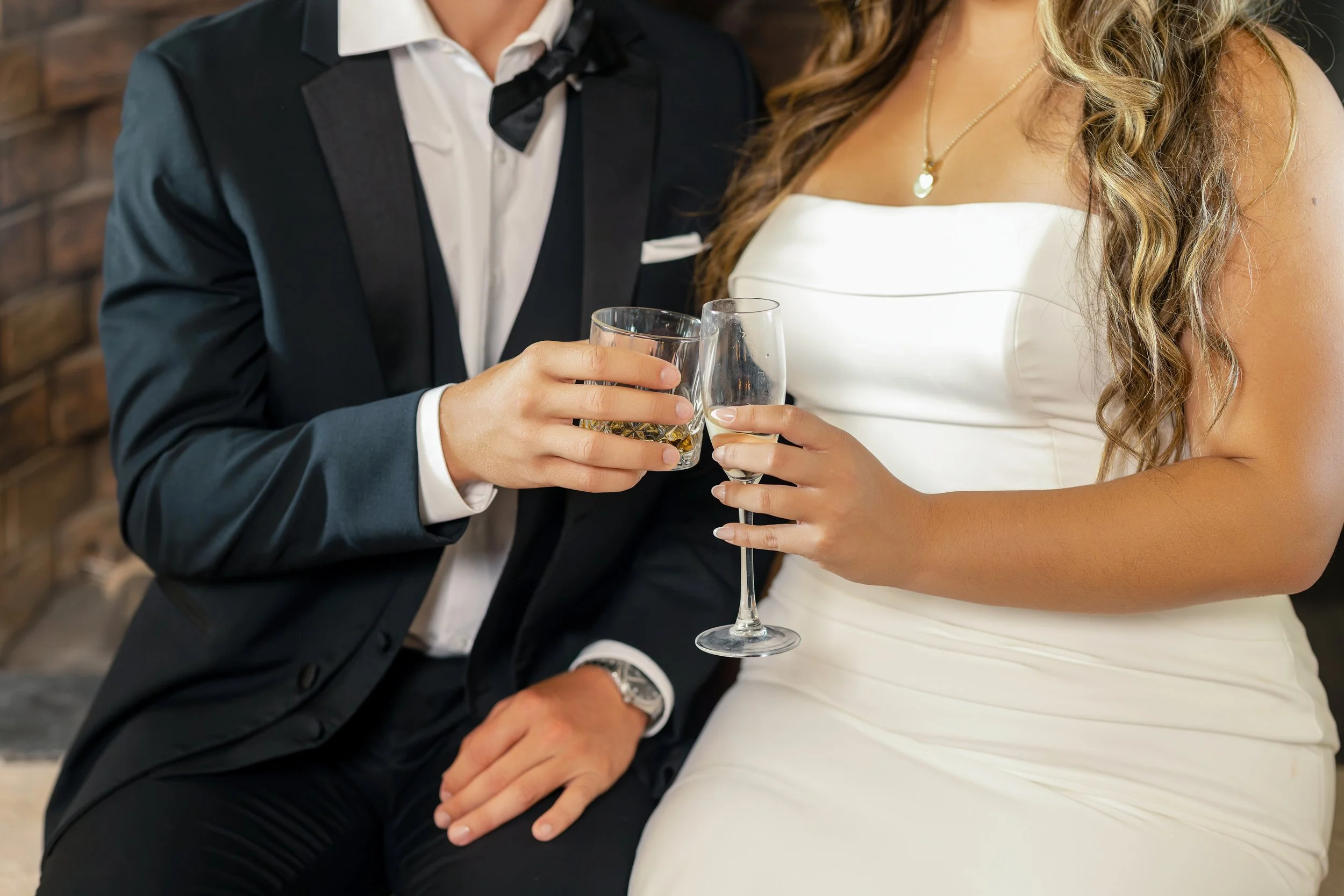 Close-up of a couple dressed in formal attire, holding glasses of champagne or cocktail, with a brick wall behind them.