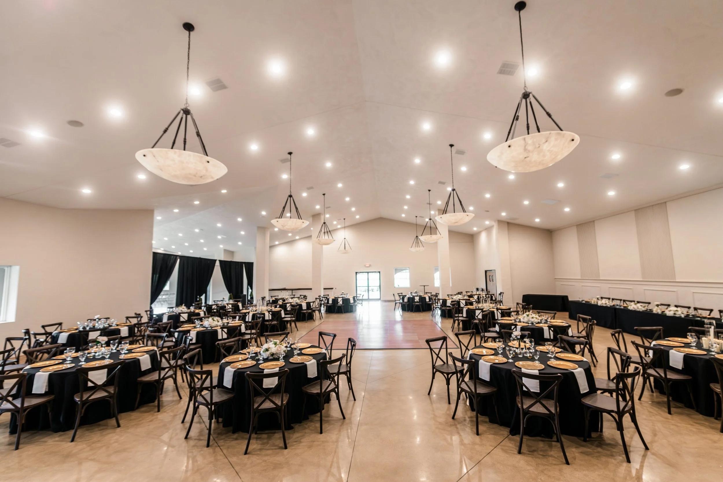 A large banquet hall with multiple round tables covered with black tablecloths, set with plates, glasses, and floral centerpieces. The hall has a high ceiling with numerous small recessed lights and large pendant lights hanging down. There are black curtains on the windows and a dance floor in the middle of the room.