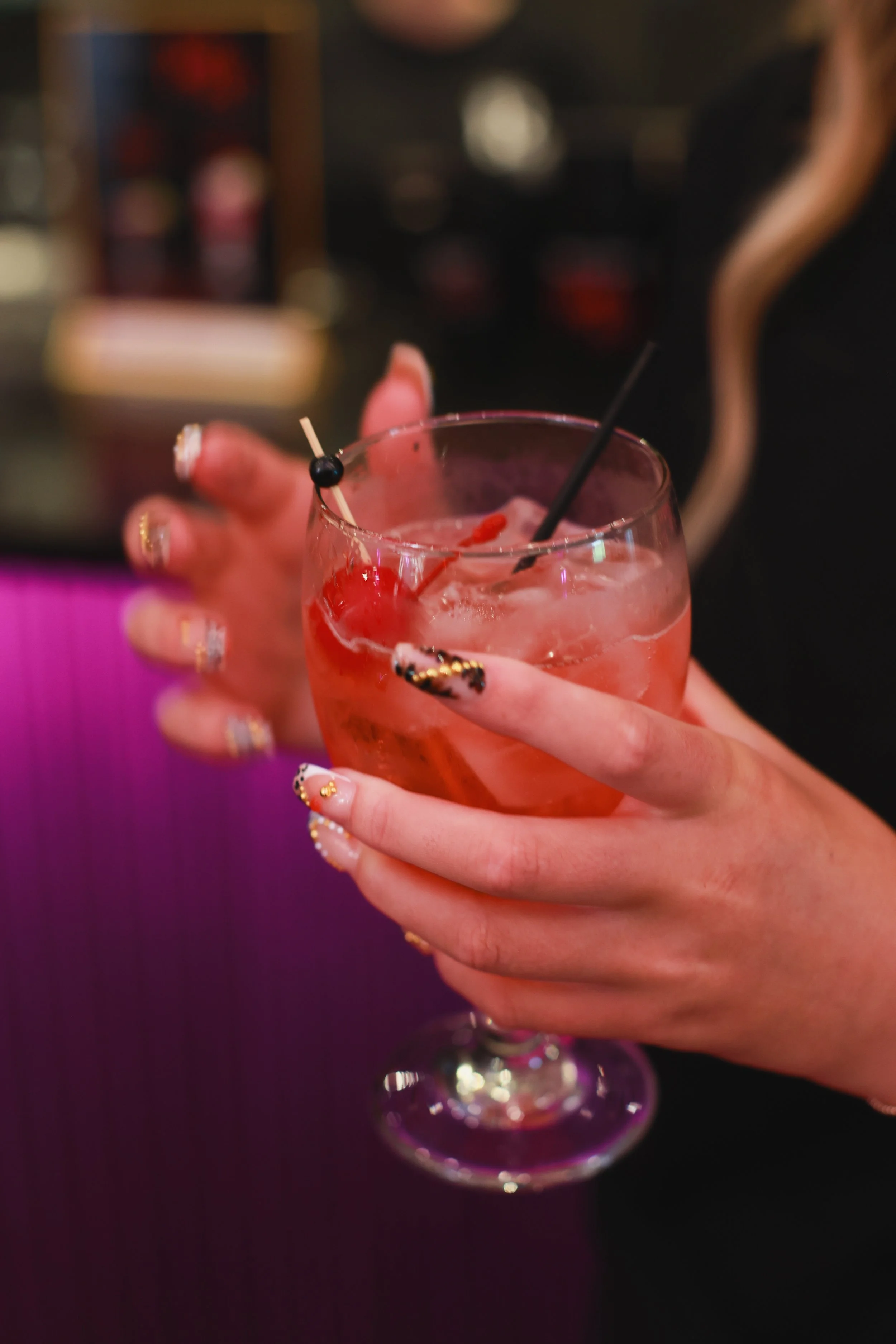 A person holding a pink cocktail in a wine glass with ice, a black straw, and decorative black berries, with a blurred background.