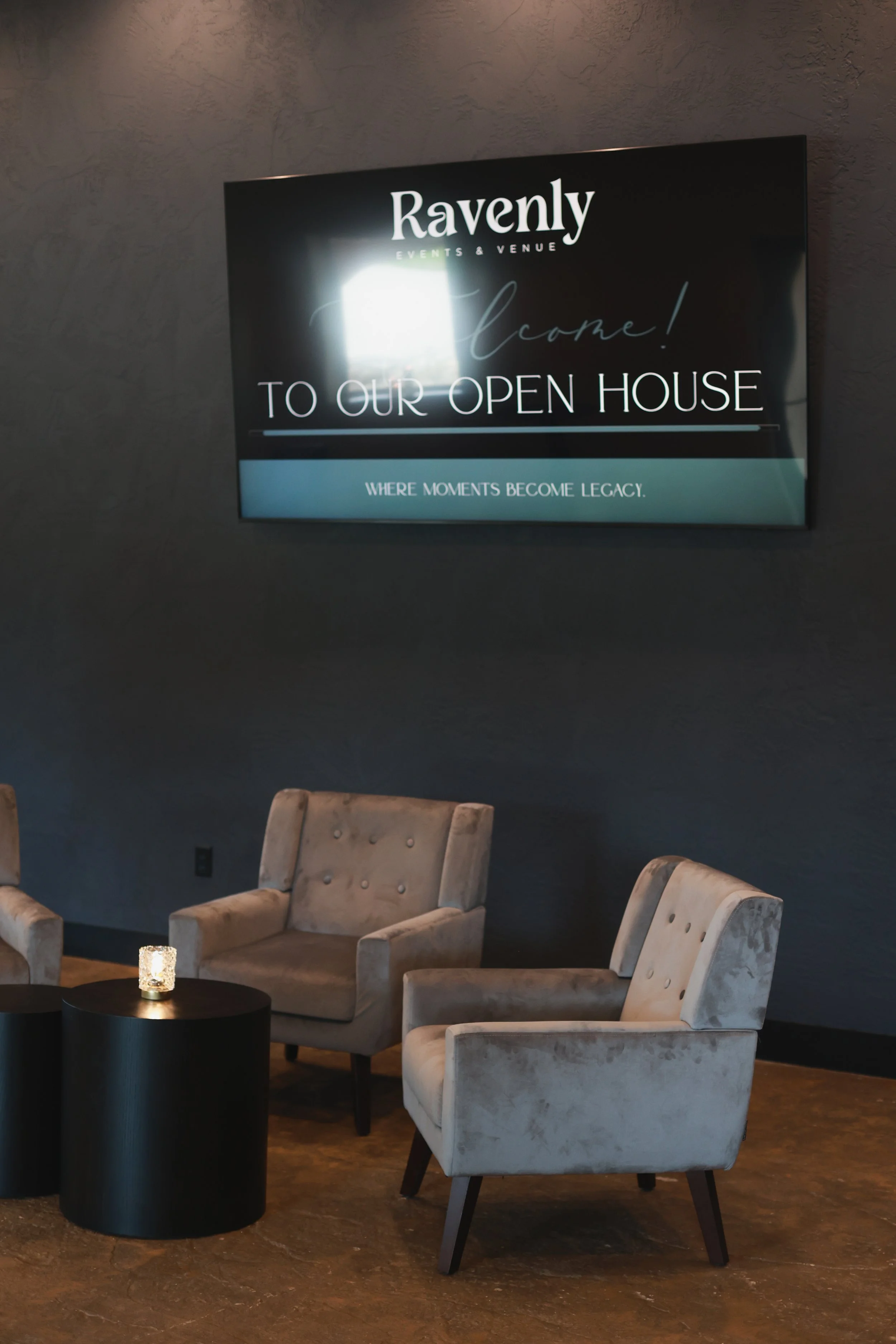 A seating area with three beige upholstered chairs with black wooden legs arranged around a small black round table with a lit candle. On the wall behind, a large flat-screen TV displays an opening message for Ravenly Events & Venue, welcoming guests