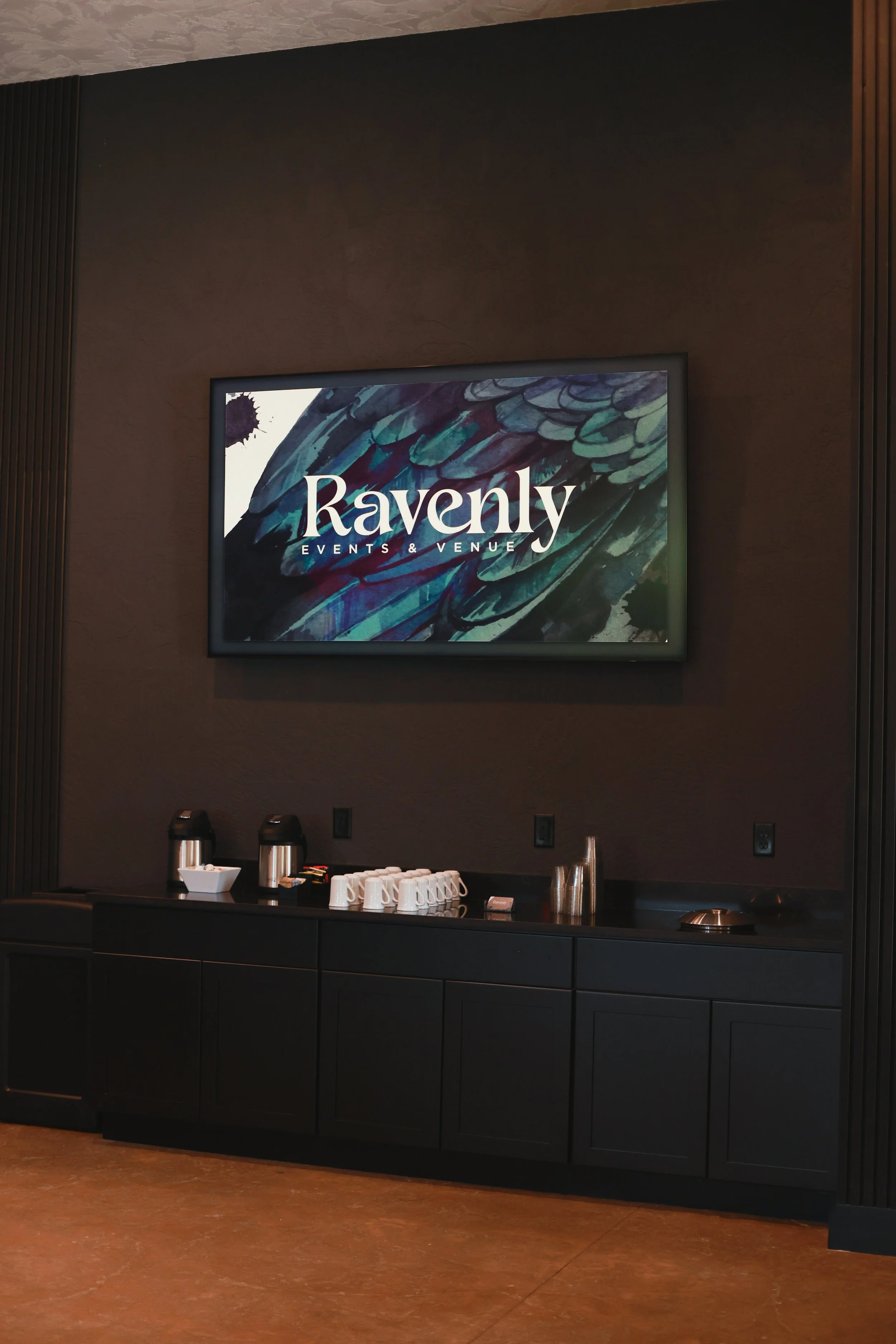 A digital display shows the logo for Ravenly Events & Venue, featuring a colorful watercolor feather design background.