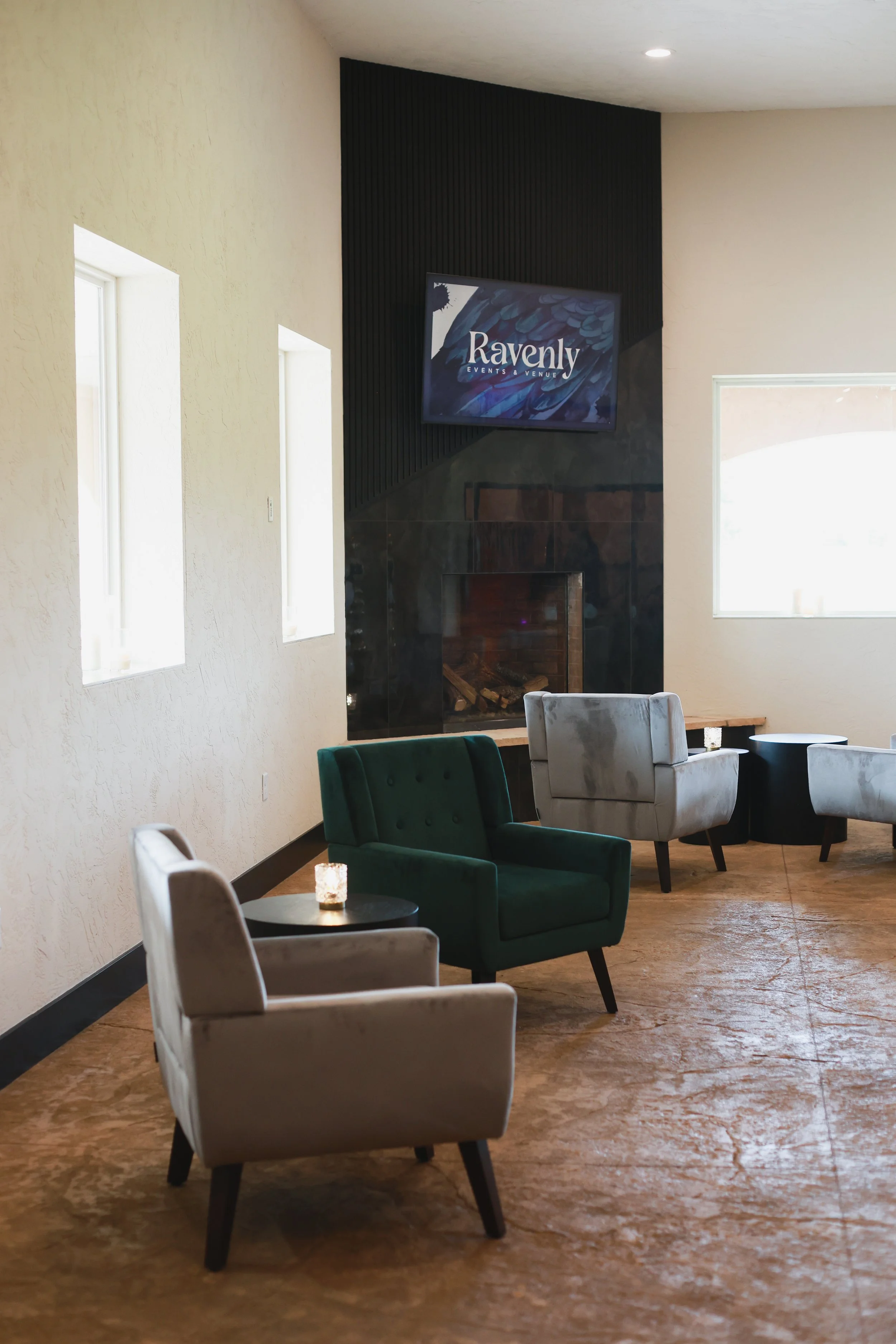 A cozy lounge with chairs around small tables and a modern fireplace with a flat-screen TV above it, displaying the text 'Ravenly Events & Venue.'