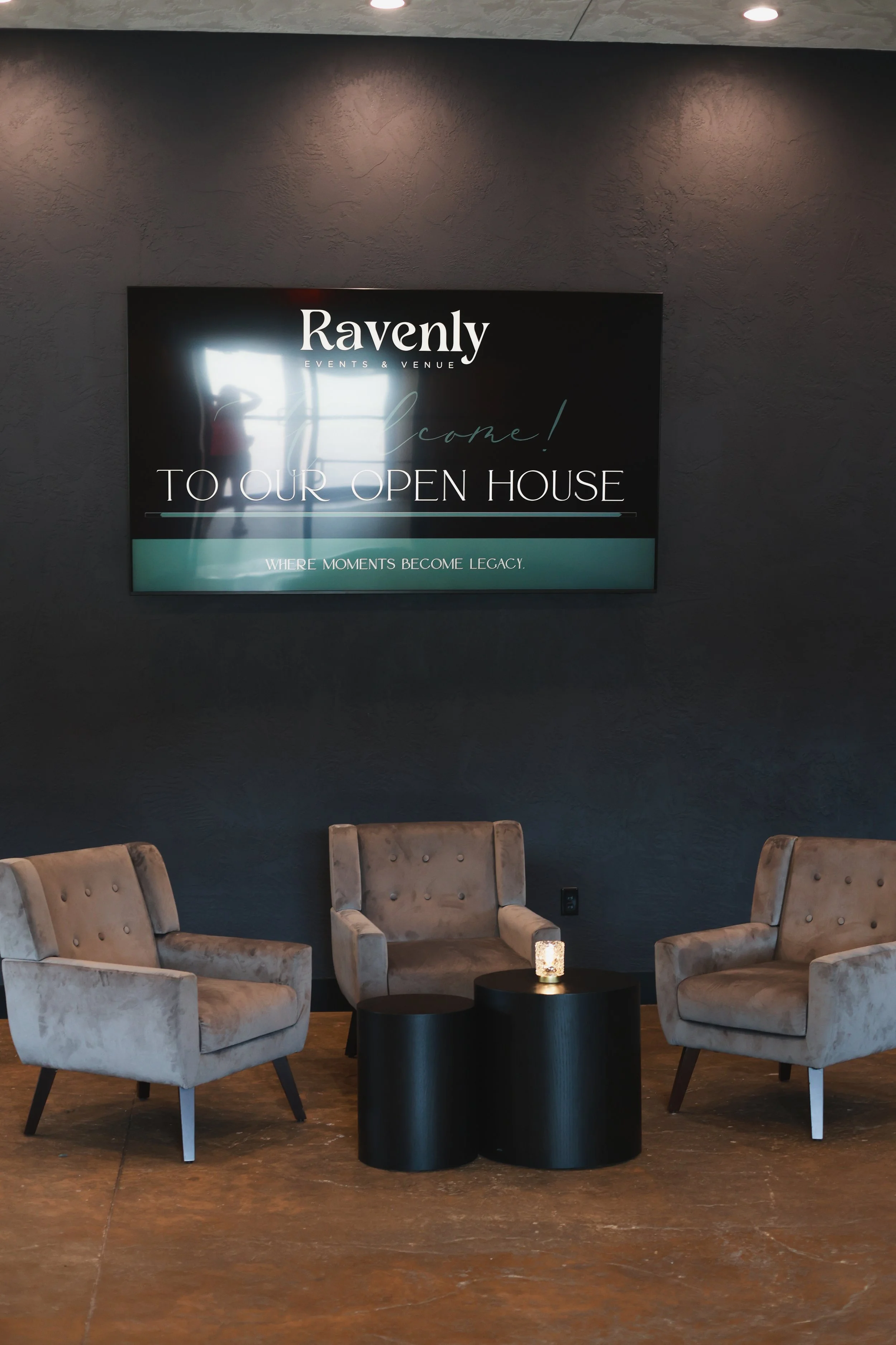 A lounge area with four beige upholstered chairs around a small black round table, a white candle, and a large screen on the dark gray wall displaying an event invitation to an open house at Ravenly, an events and venue space.