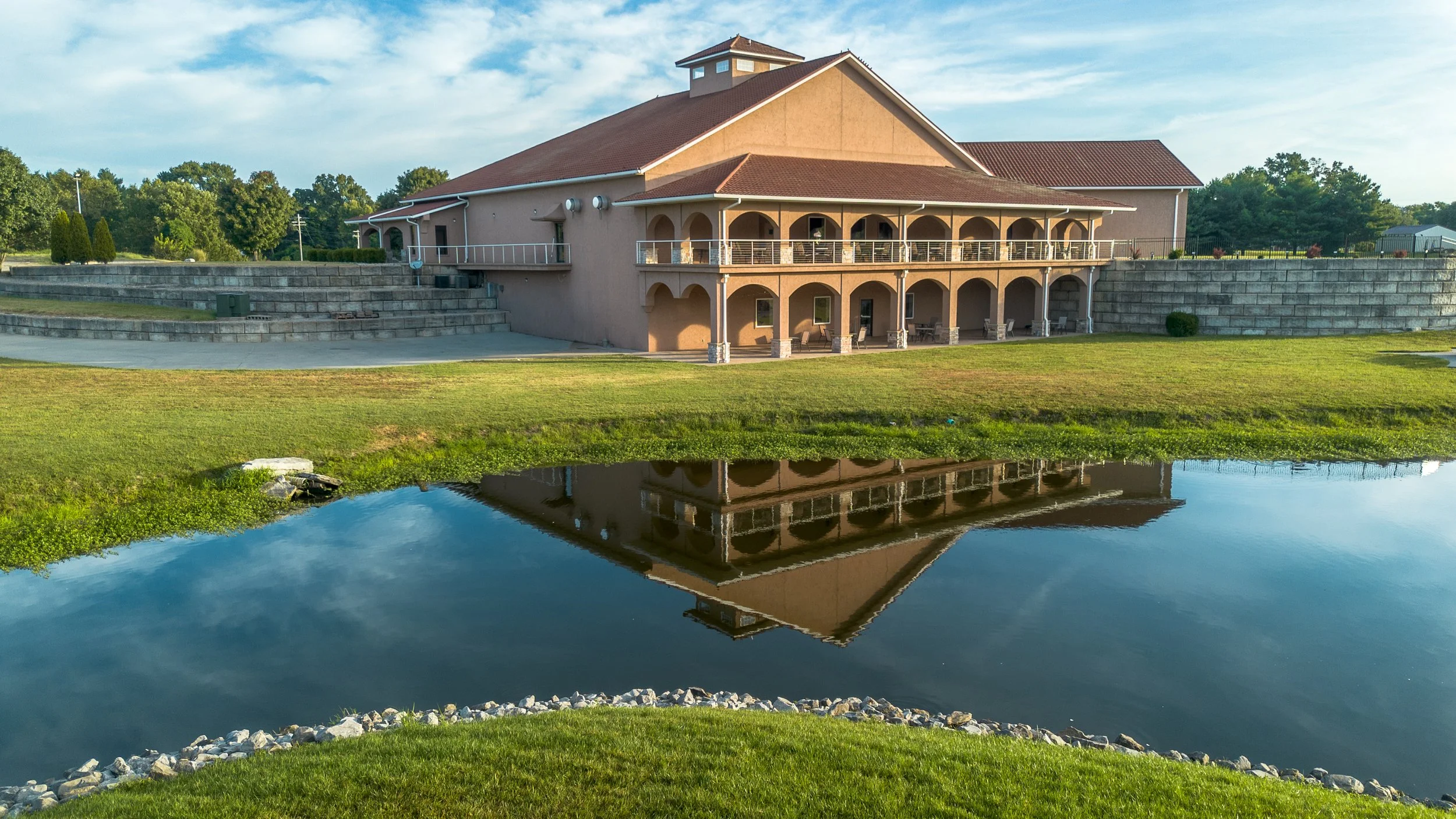 A tan building with a red roof is situated next to a small pond, with the building's reflection visible in the water. The building has a two-story porch with arches and outdoor furniture. There are trees and a grassy area surrounding the scene under a partly cloudy sky.