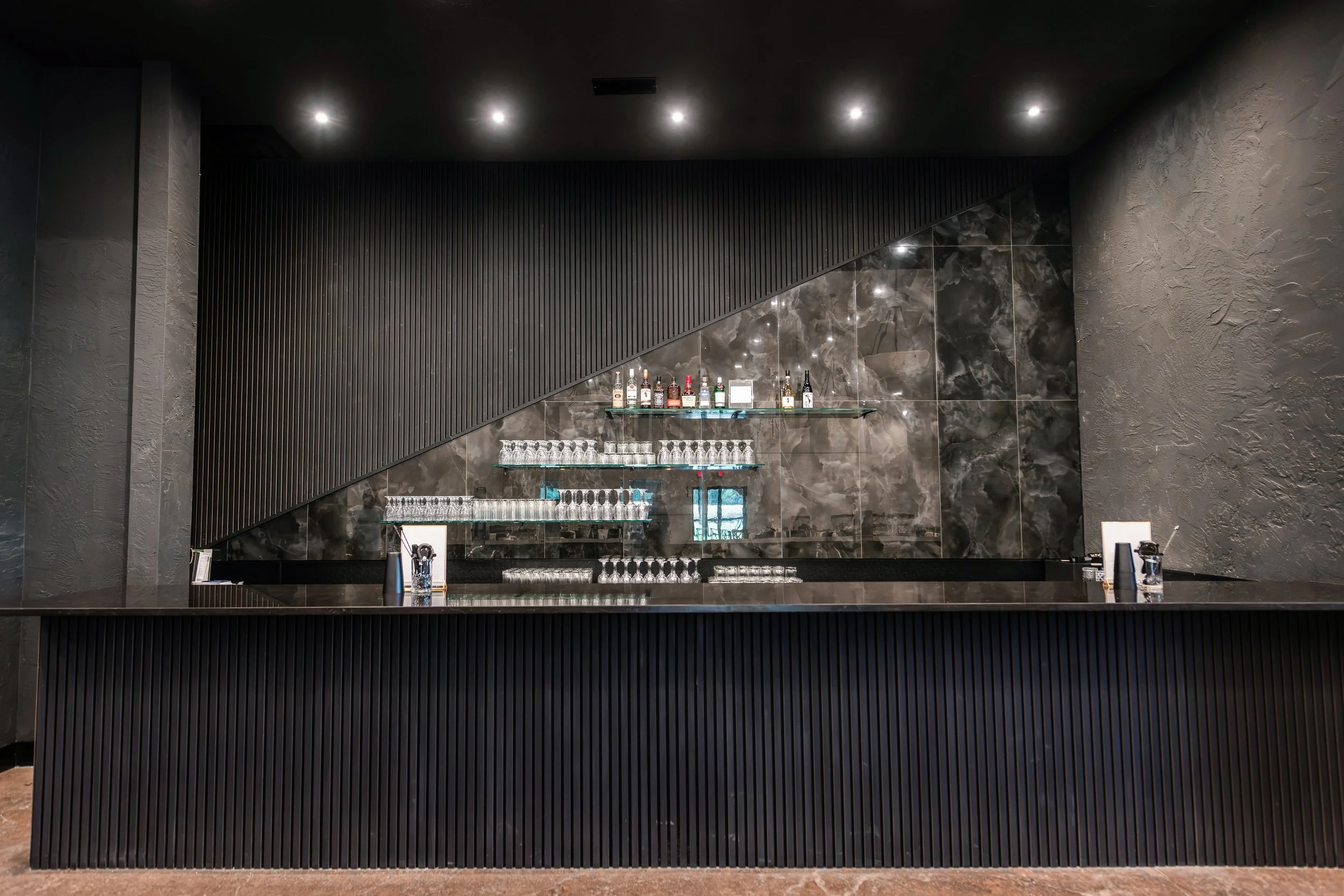 Interior of a modern bar with black textured walls, a glass shelf with liquor bottles, glassware, and bar tools, under a sloped black ceiling with recessed lighting.
