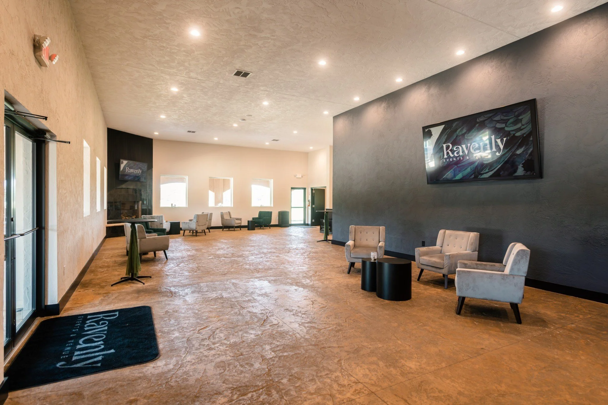 Spacious interior of an event venue with beige walls, wood flooring, multiple chairs, and a large TV screen displaying the text 'Ravenly Events & Venue'.