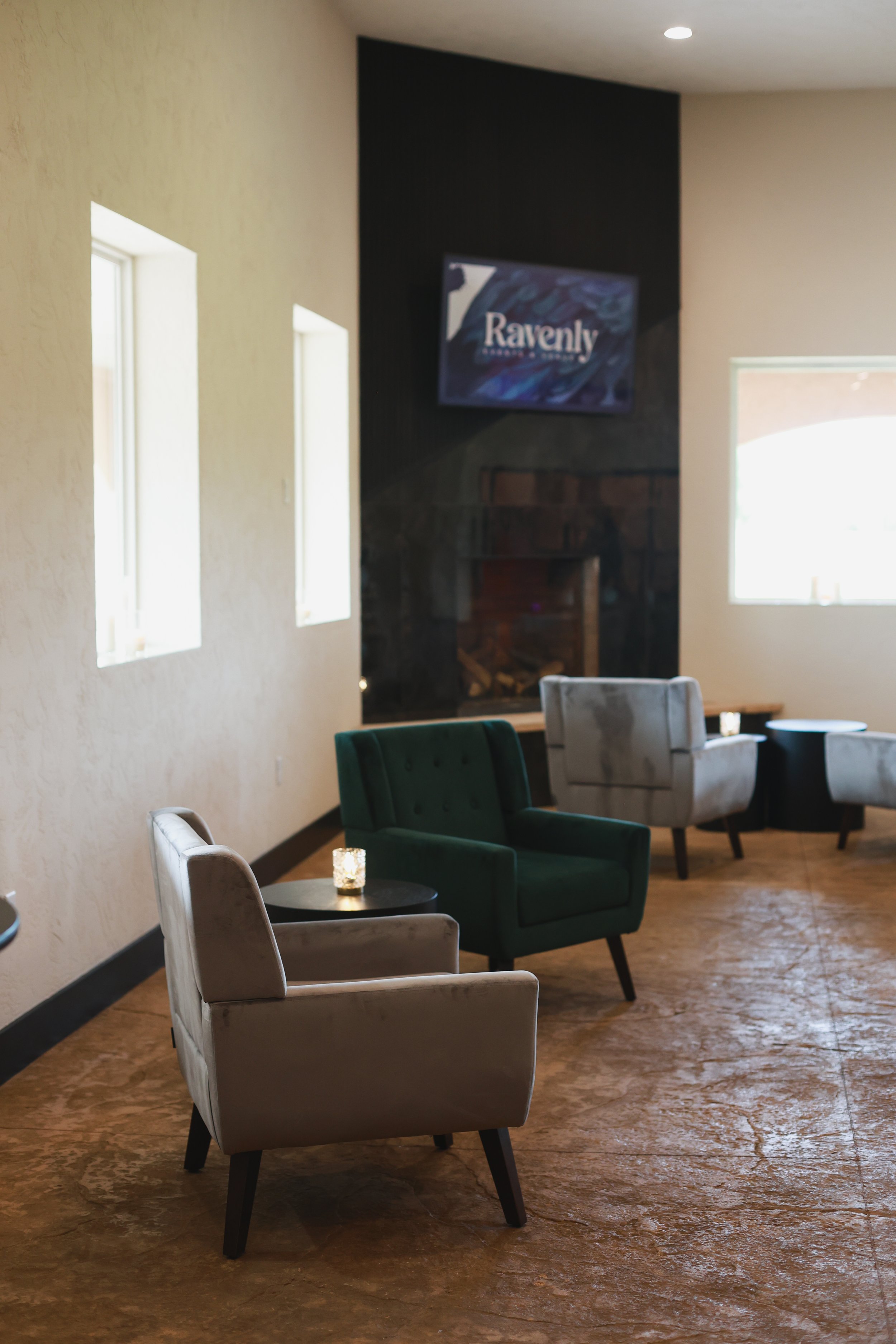 A cozy indoor lounge area with plush chairs in beige, green, and light gray, a small black side table with a candle, and a large window letting in natural light. A TV on the black accent wall displays the logo "Ravenly". There is a fireplace below th