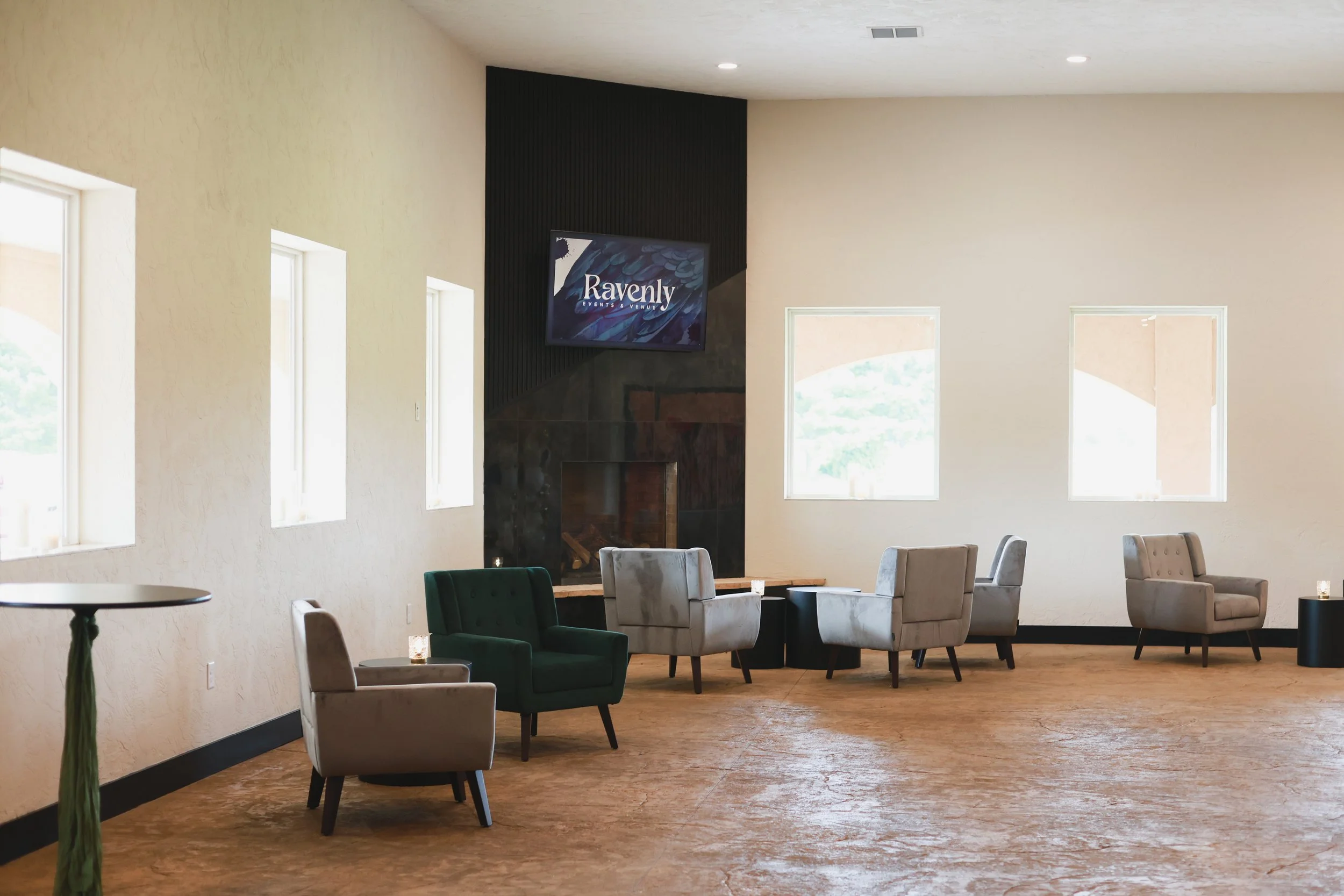 A spacious, well-lit lounge area with multiple chairs, a fireplace, windows, and a TV screen displaying the logo 'Ravenly Events & Venue'.