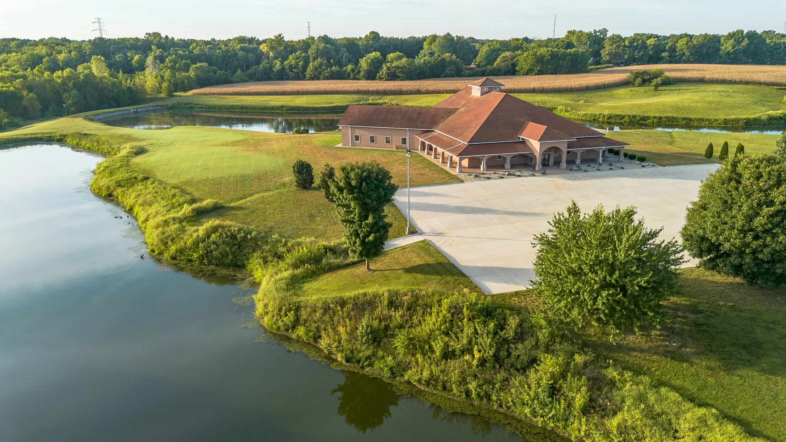 Aerial view of a building with a red roof near a pond, surrounded by lush green grass, trees, and fields.